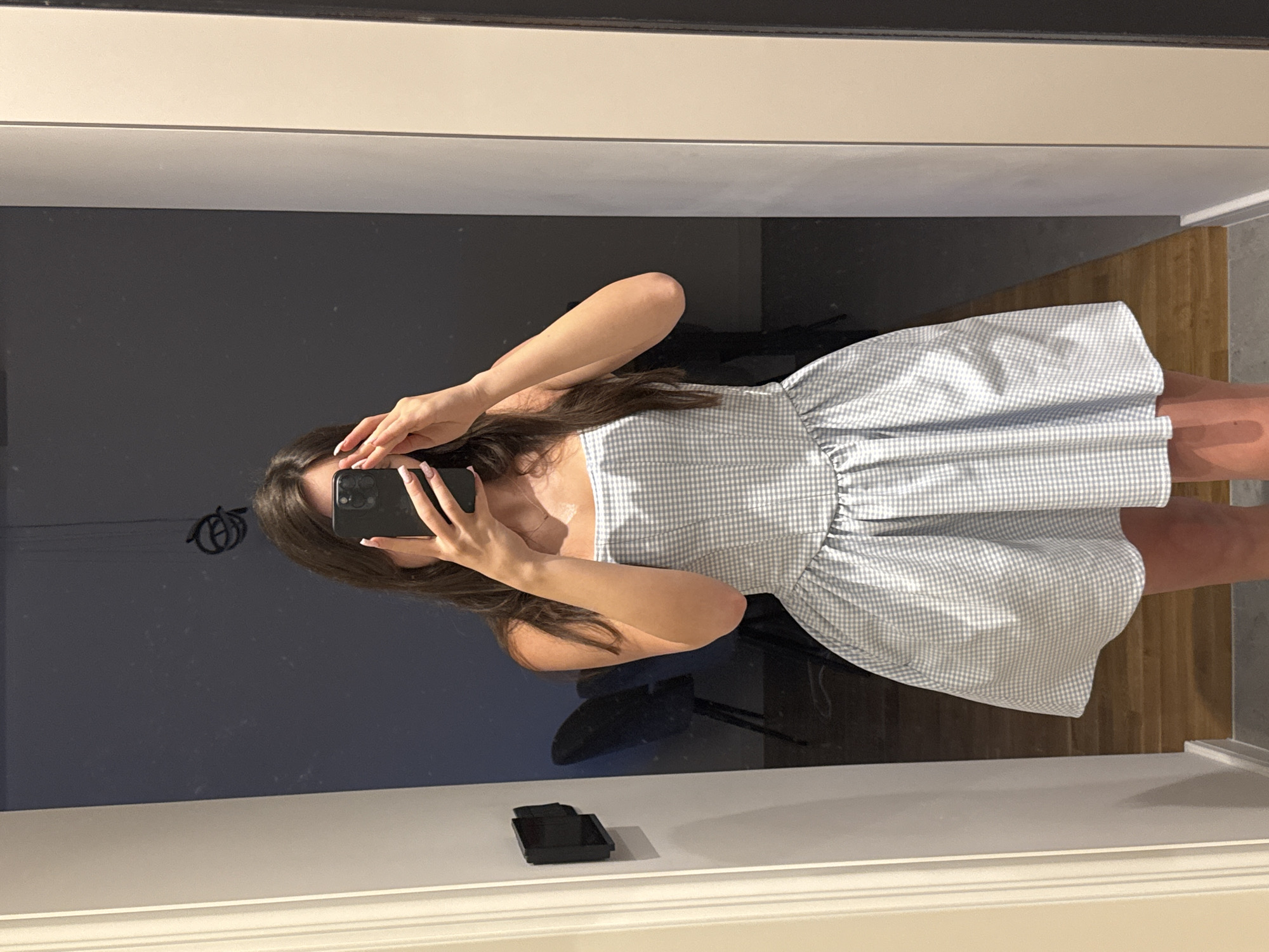 A young person is captured in a mirror selfie, holding a black smartphone that obscures their face. The image is taken from a slightly high angle. The person has long, dark hair flowing over their shoulders and is wearing a strapless or off-the-shoulder dress with a light blue and white small-checkered (gingham) pattern. The dress features a gathered or ruffled waistline that forms a full, flared skirt. Their manicured nails, painted a light pink or nude color, are visible as they grip the phone.
The reflection in the mirror shows an indoor setting. Behind the person, a dark, possibly navy or charcoal, wall is visible. Below the mirrored wall, there's a white or off-white trim or ledge, on which a small black rectangular object rests in the bottom-center of the frame. To the right side of the mirror's reflection, a wooden floor with visible planks and a light grey wall or another surface can be seen, indicating the room's layout. The lighting appears to be even and diffused, suggesting an indoor environment during daytime or well-lit conditions. No specific emotions are discernible due to the face being hidden. There is no visible text in the image.