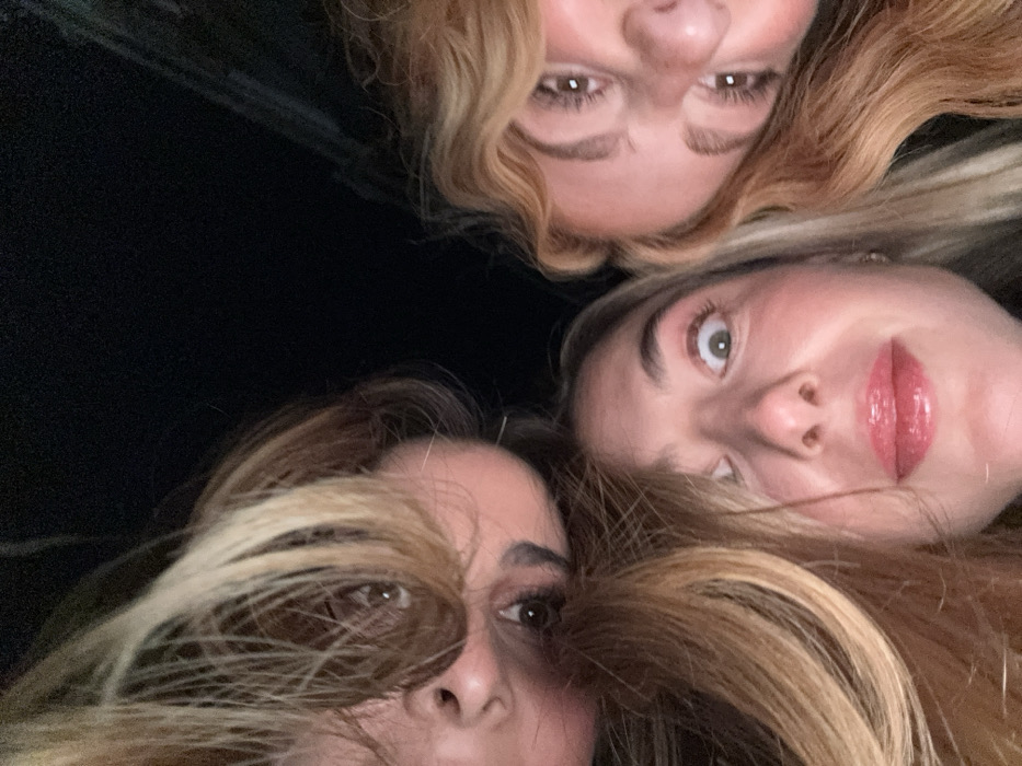 Three women cluster closely together, their faces angled towards the camera in a selfie-style shot. The women have fair skin and light-colored hair. The top woman has curly blonde hair. The woman on the right has long blonde hair and a glossy pink lip. The woman on the left has long, straight brown hair. The background is dark and nondescript, focusing attention on the trio's close proximity and facial features.