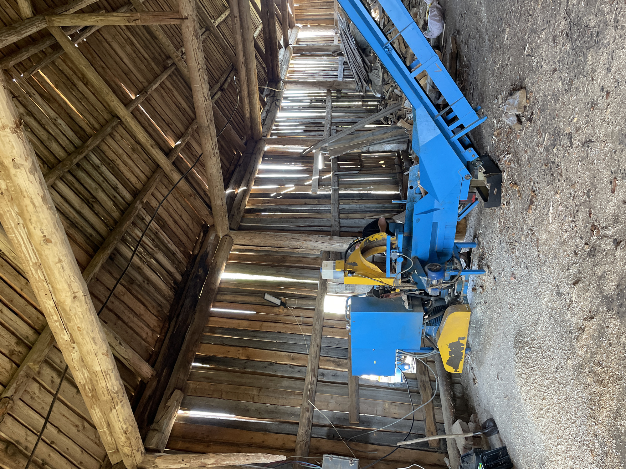 The image depicts the interior of a dilapidated wooden structure, possibly a barn or shed, in the city of Van'ki, Russia. The building's walls and roof are constructed from rough-hewn wooden planks, with gaps allowing sunlight to penetrate. A large, blue and yellow machine dominates the foreground, its purpose unclear. The ground is covered in a mix of dirt, debris, and wood shavings. No people are visible in the image. The overall atmosphere is one of disrepair and neglect.