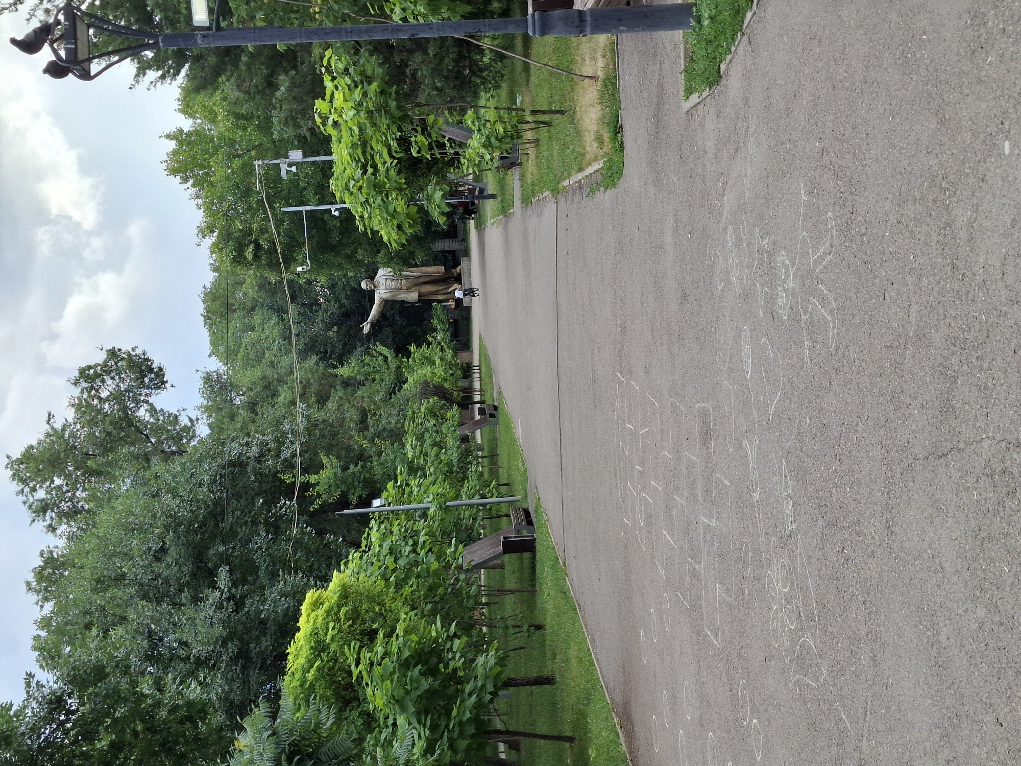 This media file depicts a park scene in Almaty, Kazakhstan. The foreground is dominated by a paved pathway with chalk drawings of hopscotch squares and other playful figures, suggesting children have been active here. Lush green trees and foliage line the pathway, indicating a pleasant, likely warm, season. Several park benches are visible, interspersed among the trees and along the path, offering places for rest and contemplation.
Further down the path, a prominent statue of a man in a suit, with one arm outstretched, stands amidst the greenery. The statue is positioned to draw the viewer's eye into the depth of the park. In the background, dense trees create a verdant backdrop, with some sky peeking through the canopy. Overhead, a lamppost with what appears to be security cameras attached is visible, along with wires crisscrossing the scene, suggesting park infrastructure.
The sky is partly cloudy, with patches of white clouds against a lighter sky, hinting at a day that is not entirely sunny but still bright. The overall atmosphere is serene and invites a leisurely stroll through the park. The specific time of day is difficult to ascertain precisely, but the lighting suggests it is daytime. The presence of the statue and the general layout of the park are consistent with public spaces often found in Almaty. No legible text is present in the image.
