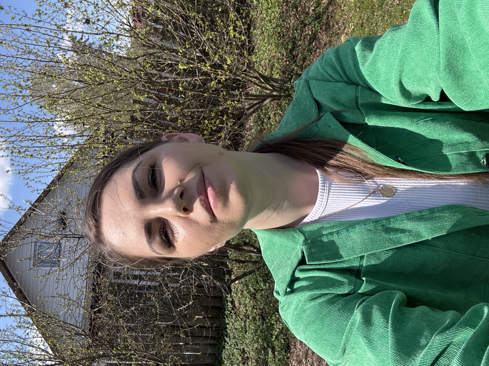 A young woman poses for a selfie outdoors in Razdolie, Russia. She wears a white tank top and a green corduroy jacket. Behind her is a wooden building with a window, partially obscured by bare tree branches. There is green vegetation and grass in the background. The sky is blue with a few clouds. The lighting suggests it is daytime.
