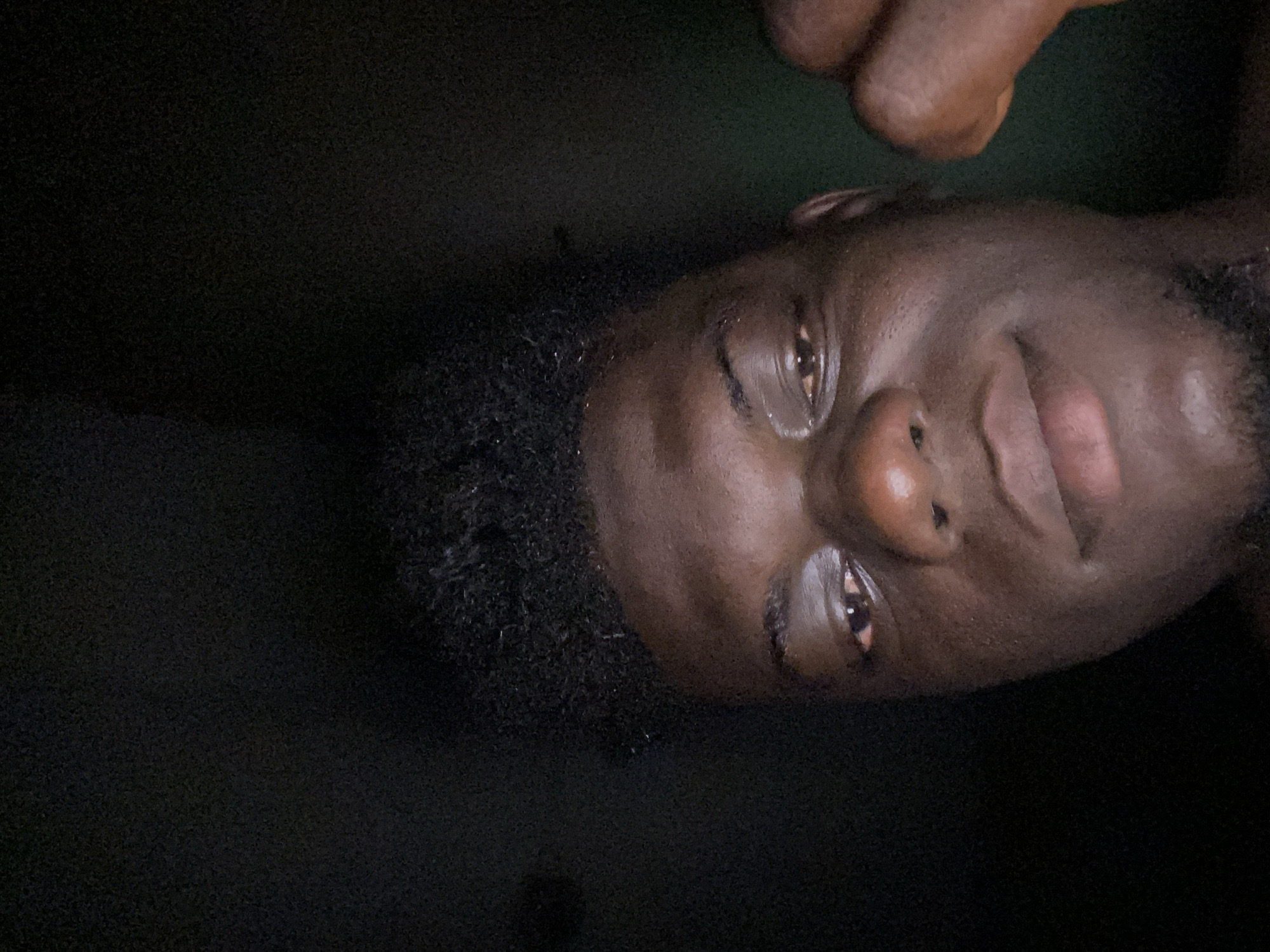 The image is a close-up of a dark-skinned man's face. He has short, dark, curly hair and appears to be in low lighting or darkness. There are fingers above his head. The location is unknown.