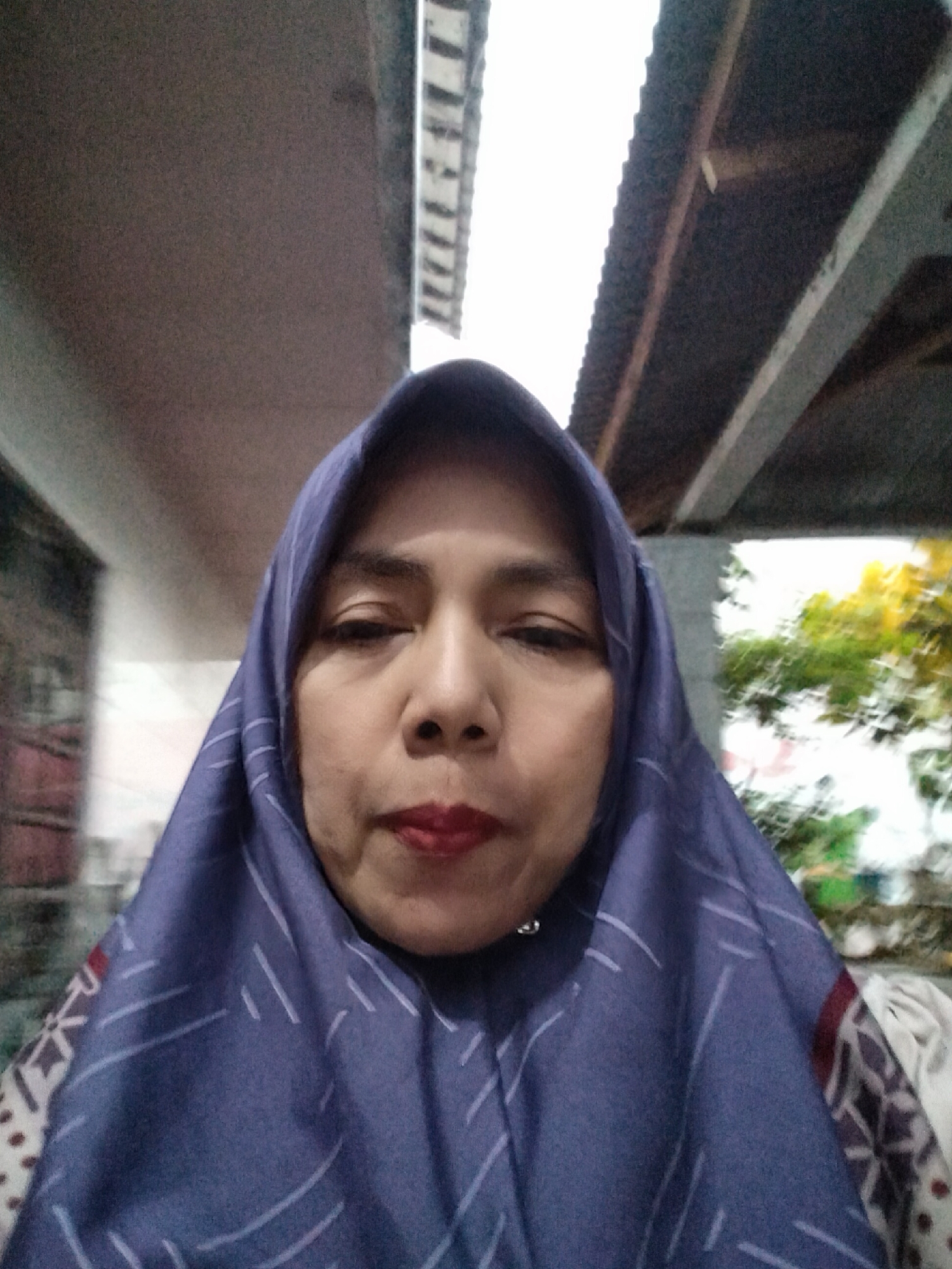 A close-up, selfie-style, medium shot captures a woman's face and upper body. She is wearing a purple hijab adorned with a white geometric pattern. Her face is turned slightly towards the camera, with her lips accentuated by red lipstick. The background suggests an outdoor setting, possibly near a building with a corrugated roof, and some greenery is visible in the blurred distance. The lighting is somewhat dim, but the woman's face is well-lit, indicating the use of a flash or a strong light source. The image does not provide specific landmarks or visual cues that would definitively place the location within Sukamanah, Indonesia, beyond what is stated in the prompt.