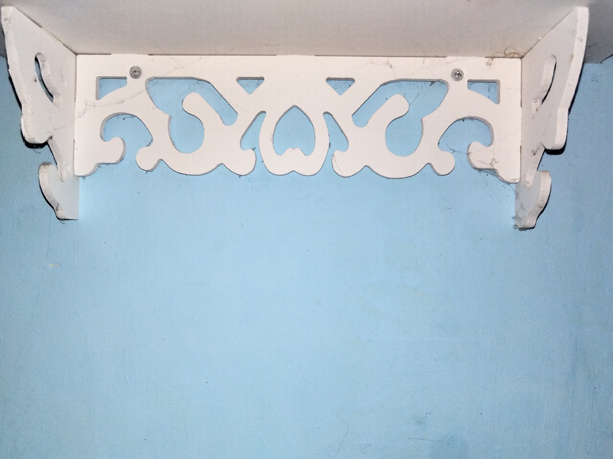 This media file shows a white decorative shelf bracket mounted on a light blue wall. The bracket has an ornate design with cutouts resembling hearts and curves. Two screws are visible securing the bracket to the shelf above. The wall appears to be painted plaster and shows some texture and minor imperfections. There are wisps of cobweb clinging to the bracket and shelf, suggesting the area may not be frequently cleaned. The lighting is even, indicating an indoor setting, likely a room. There is no visible text in the image. The scene is static, with no activity taking place. The overall impression is one of a simple, decorative home furnishing against a plain wall. The location context of Kualaraja, Indonesia, does not offer specific visual cues within the image itself.