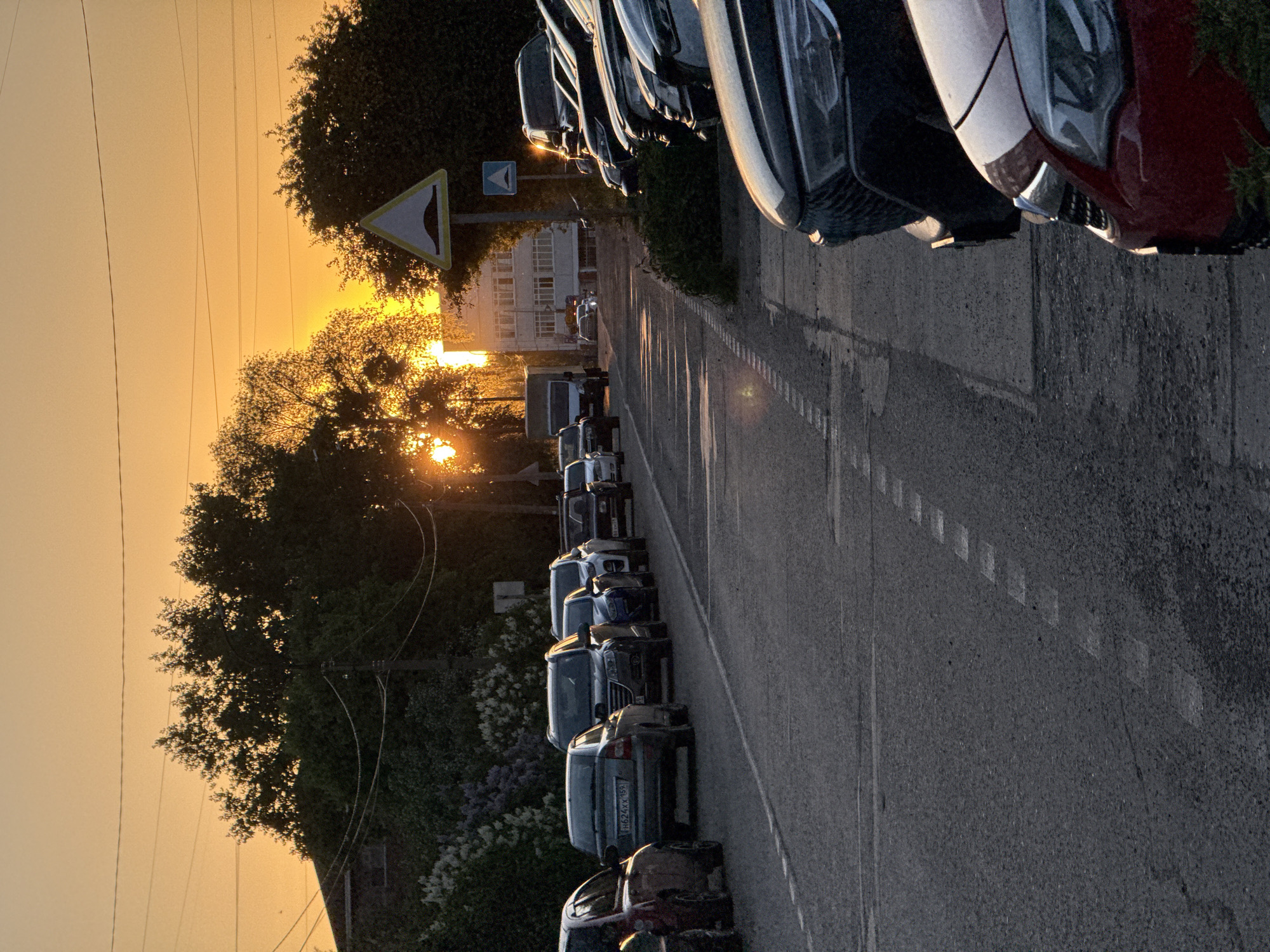 The image depicts a street in Chaikovsky, Russia at sunset. Cars are parked along both sides of the road. A yield sign is visible on the left side of the road, next to a tree. A building is visible in the background. The sun is setting in the distance, creating a golden glow.