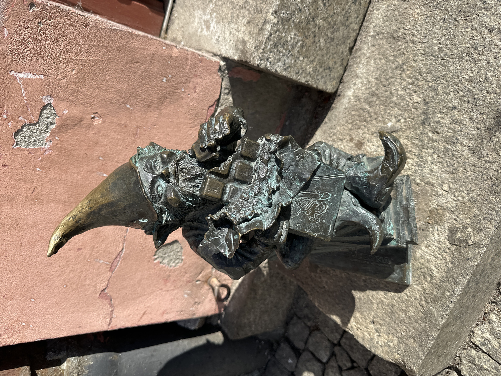 A small, bronze dwarf statue is affixed to a wall and sidewalk in Wrocław, Poland. The statue wears a tall, pointed hat and is sitting with an open book. The statue is situated where a pinkish wall meets a gray stone sidewalk. The dwarf appears to be reading, or perhaps even writing something. The statue is likely one of the city's numerous dwarf statues, which are a popular tourist attraction. The background consists of the textures of the stone and plaster.