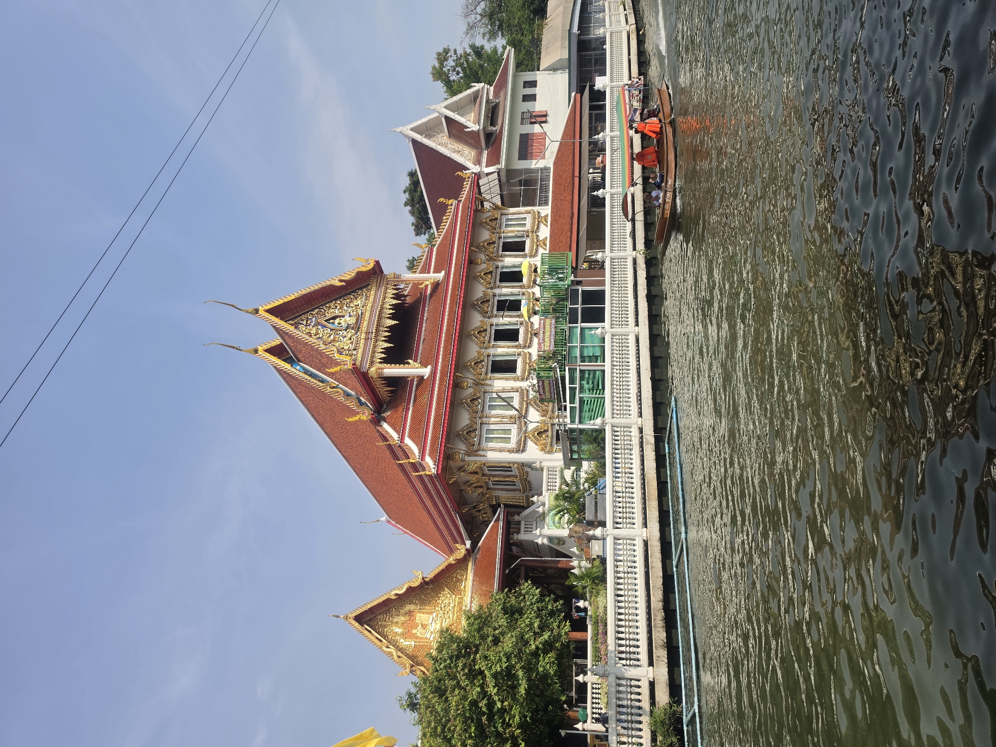 The image depicts a waterfront scene in Bangkok, Thailand, featuring a traditional Thai temple and a narrow canal with a longtail boat.
**Scene:** The foreground is dominated by a canal with shimmering, rippled water reflecting the sunlight. Adjacent to the canal is a temple complex, characterized by its ornate architecture, multi-tiered roofs adorned with intricate gold decorations, and a mix of red and gold color schemes. The temple appears to be built close to the water's edge, with a white balustrade separating the building from the canal.
**People:** A longtail boat is visible in the canal, containing several individuals, likely passengers, wearing orange life jackets.
**Objects:** The prominent objects in the scene are the temple building, the canal, the longtail boat, and a white fence running along the water. There are also trees visible in the background. The image shows powerlines stretched high above.
**Environment:** The environment suggests a sunny day, with a clear blue sky visible above the temple. The canal setting points to a waterway often used for transportation or recreation in Bangkok.