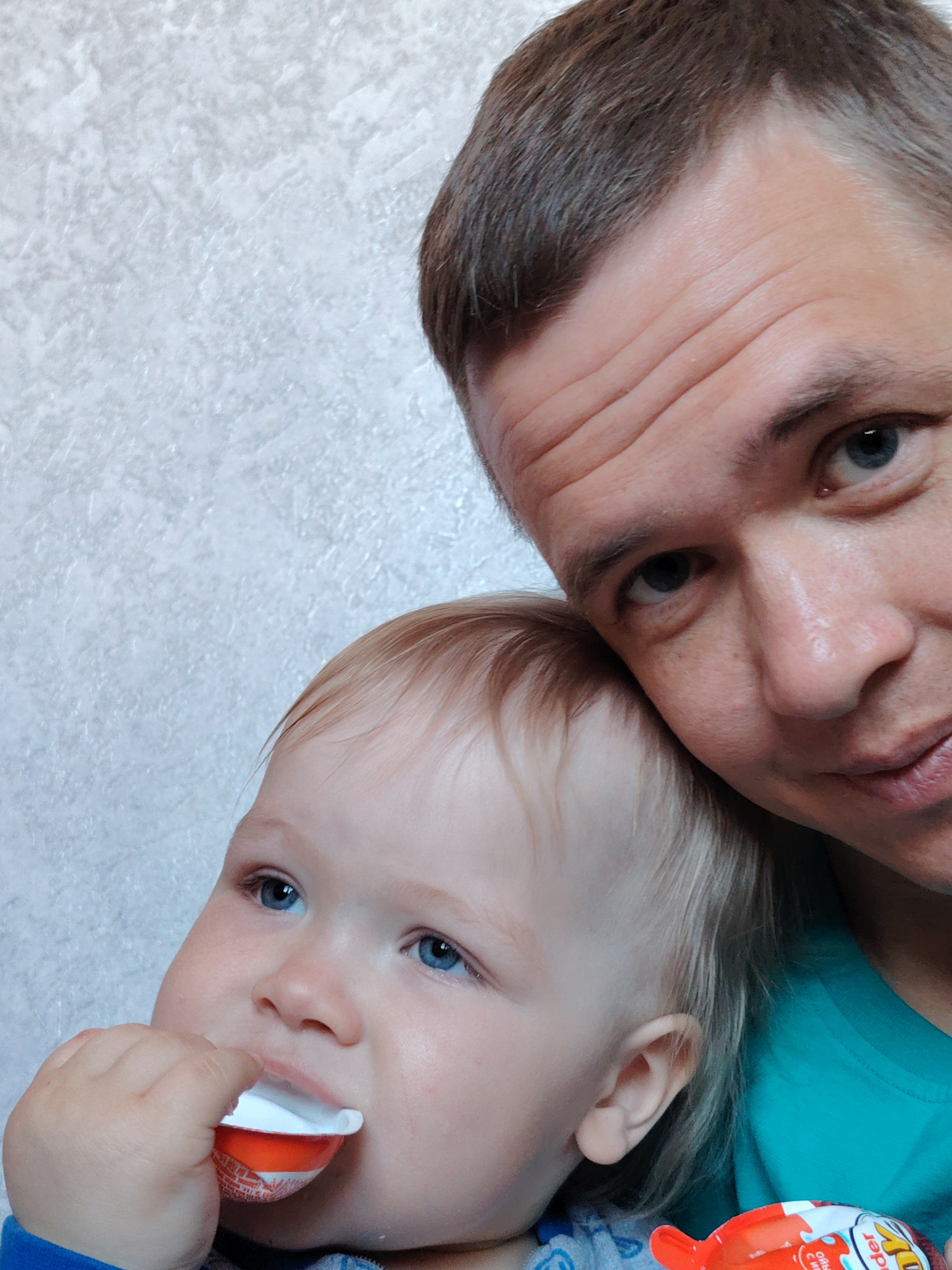 A close-up selfie shows a man and a young child indoors, likely in Chaikovsky, Russia. The man, positioned in the upper right, has short light brown hair, blue eyes, and visible lines on his forehead, looking towards the camera with a subtle smile. He wears a teal shirt. His head is gently resting against the child's, conveying a tender connection.
The child, a toddler with light blond hair and bright blue eyes, occupies the lower left. They are focused on a red and white Kinder Joy egg, holding a small white cup from it to their mouth as if eating. The packaging clearly displays "Kinder Joy" in white text, and additional Cyrillic text is visible on the red portion, including "ИГРУШКА" (Toy) and phrases like "ВКУСНЫЙ МОЛОЧНЫЙ ШОКОЛАД" (Tasty Milk Chocolate), indicating Russian packaging.
The background is a light-colored, textured wall, providing a simple and consistent setting. The lighting suggests an indoor, daytime environment. The image captures a candid, affectionate moment between the two individuals.