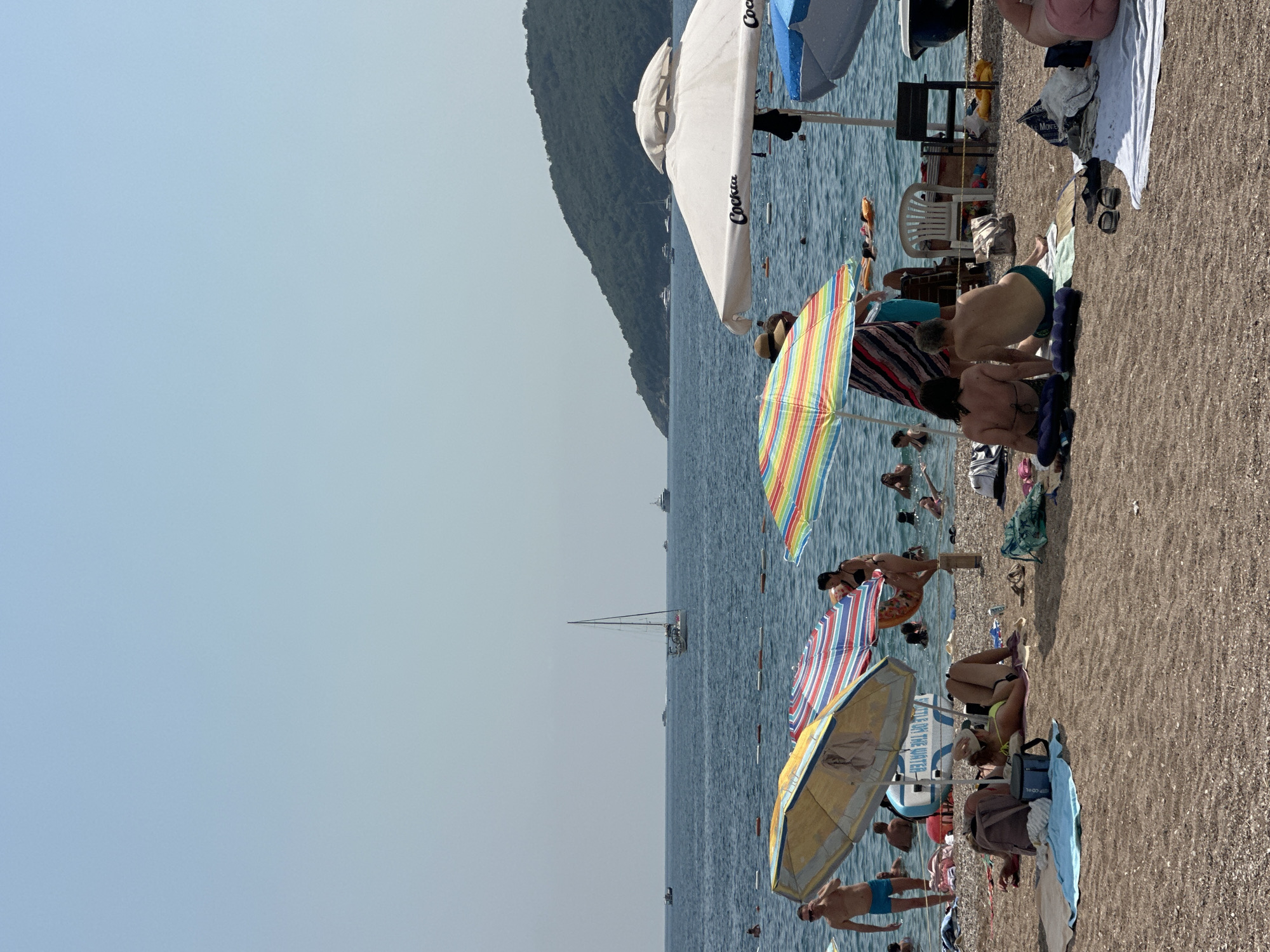 The image depicts a crowded beach scene in Budva, Montenegro, under a clear, pale blue sky. The foreground is dominated by a sandy beach with numerous people relaxing and sunbathing. Several beach umbrellas, some colorful and striped, others plain white, offer shade. Beach towels, bags, and footwear are scattered across the sand.
In the middle ground, the turquoise sea is filled with swimmers and people enjoying various water activities. A few inflatable floats are visible. The water is demarcated by a rope barrier in some sections. Several boats, including a prominent sailboat with white sails, are sailing on the horizon.
The background features a densely forested, green hill that slopes down towards the sea, characteristic of the Budva Riviera. The overall atmosphere is one of a lively summer day at the beach.