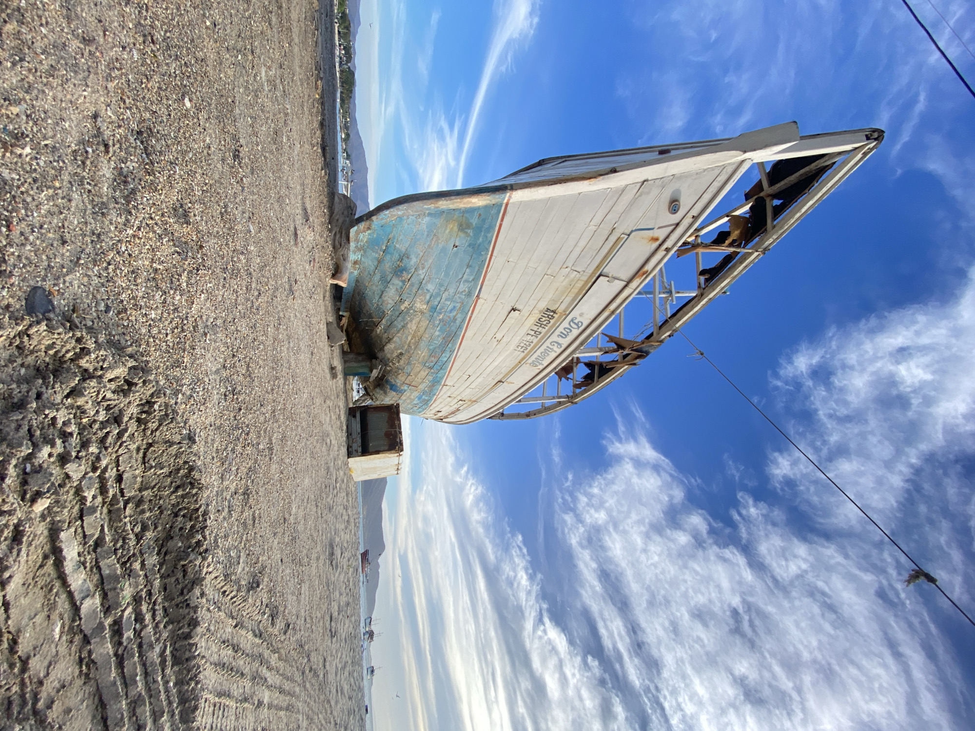 The image shows an old, derelict boat hull resting on a coarse sandy beach against a backdrop of a bright blue sky with wispy white clouds. The boat, primarily white and light blue, is heavily weathered and shows signs of decay. Its wooden frame is partially exposed, indicating significant structural damage. Writing is visible on the side of the boat, though difficult to decipher. The beach consists of mixed-size gravel and sand, and tire tracks suggest vehicle activity. The presence of other distant boats and buildings on the shoreline suggests a coastal or harbor area. It is in Juan Griego, Venezuela.