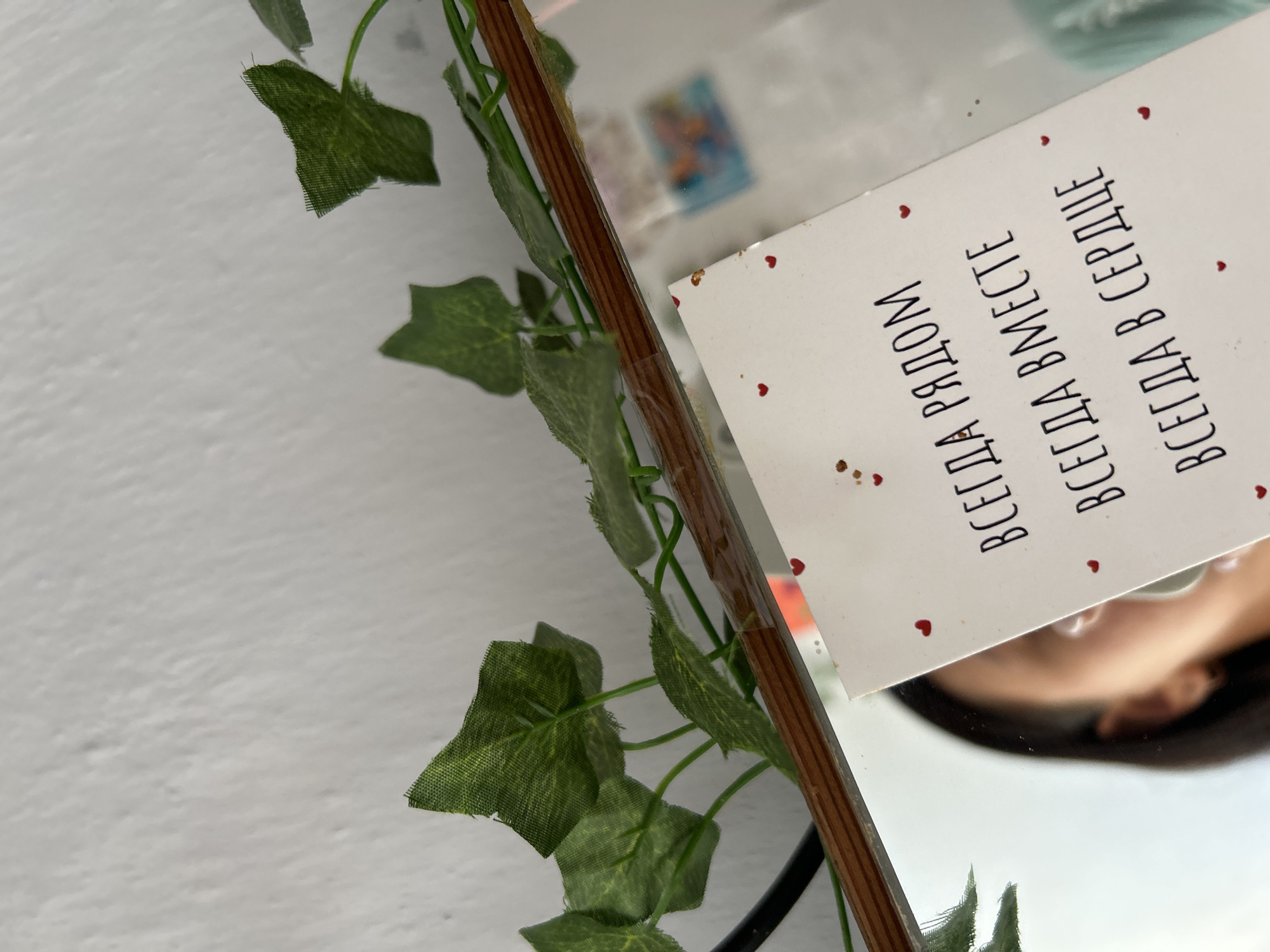 The image shows a close-up view of a white card with text written in black Cyrillic letters and small red hearts. The card is placed in front of a mirror, which reflects the left side of a person's face, including their eye, nose, and ear. The text on the card appears to be a handwritten message or quote. Artificial green leaves are visible in the foreground, arranged on what appears to be a wooden frame or shelf, which is also reflected in the mirror. The background is a textured white wall. The image composition suggests an indoor setting, possibly a personal space or a decorative arrangement. There is no visible information to indicate the location in Kerch, Ukraine.