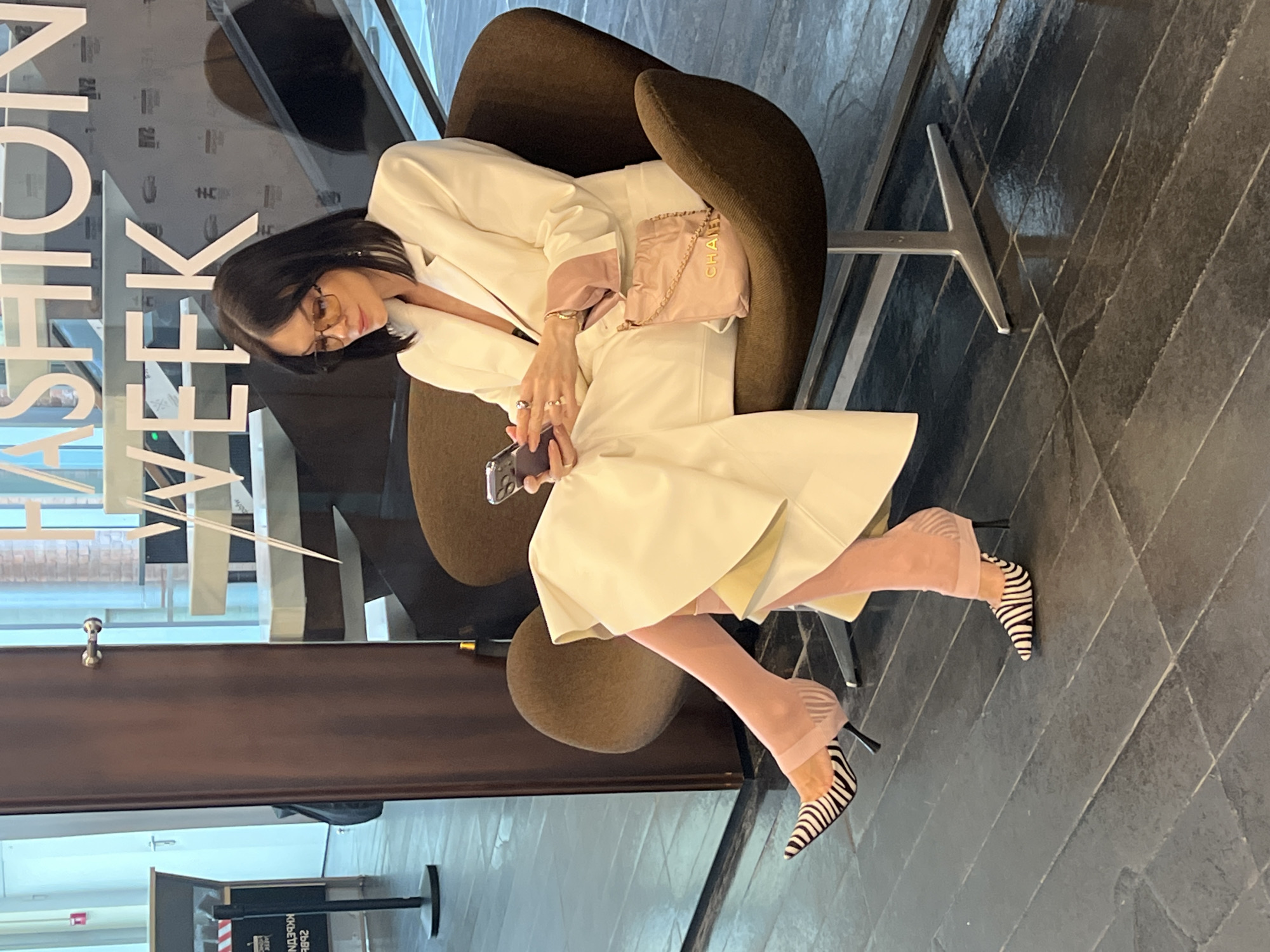 A woman is seated in a brown swivel chair at a fashion event. She is wearing a white pantsuit with pink accents, pink stockings, and zebra-striped high heels. She is holding a smartphone. A pink Chanel purse is on the chair next to her. Behind her, a sign reads "Fashion Week". The event appears to be taking place in a modern building with large windows. The building is located in Saint Petersburg, Russia. The floor is dark gray tile.