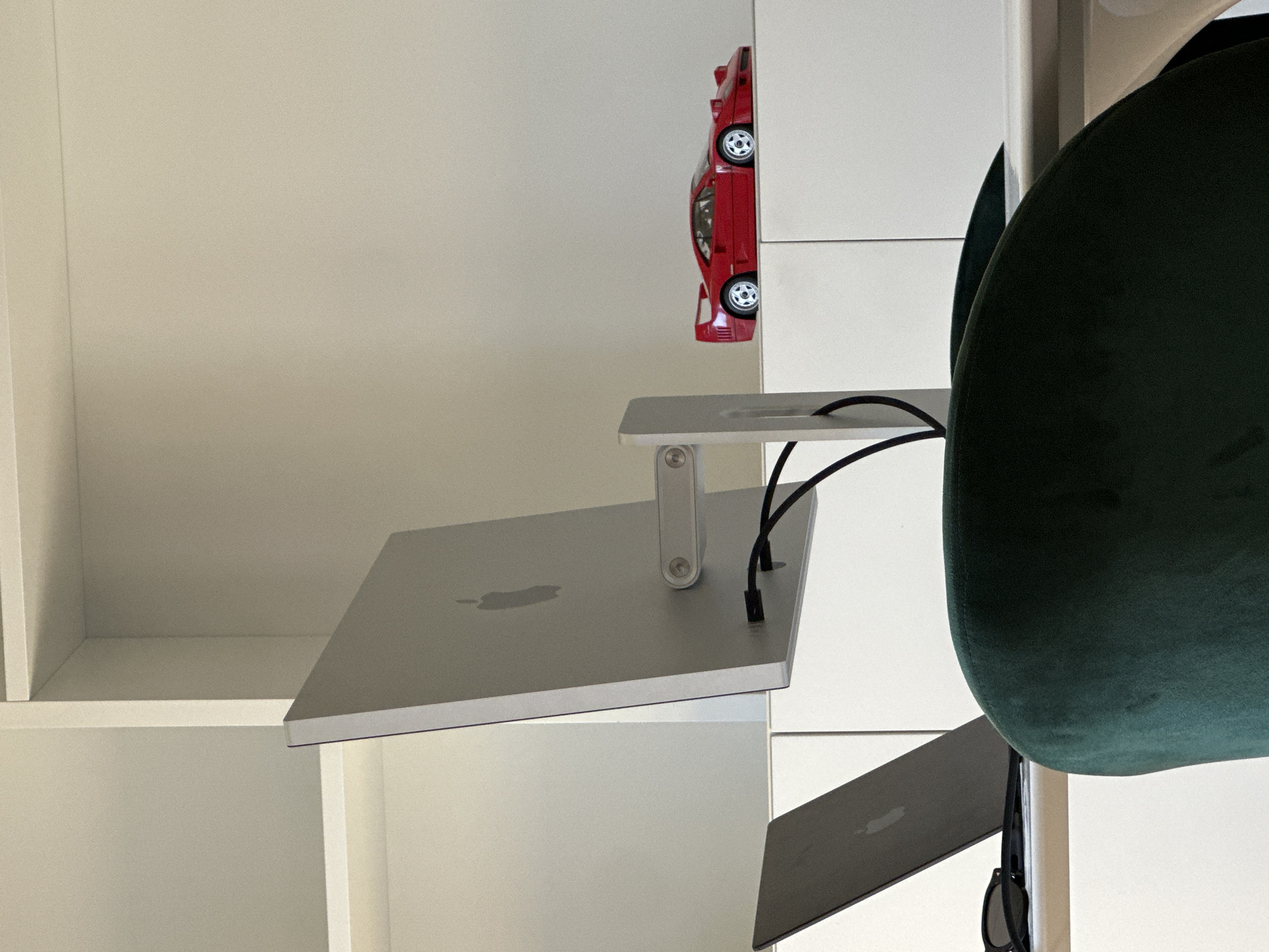 The image shows a modern, minimalist interior. There's a white shelving unit, a silver Apple iMac on an adjustable stand with cables, a red toy car positioned on the shelving, and a green upholstered chair. Sunglasses are hanging from the chair. Location is not clear based on the image, and I cannot say with certainty it is in Milan, Italy.