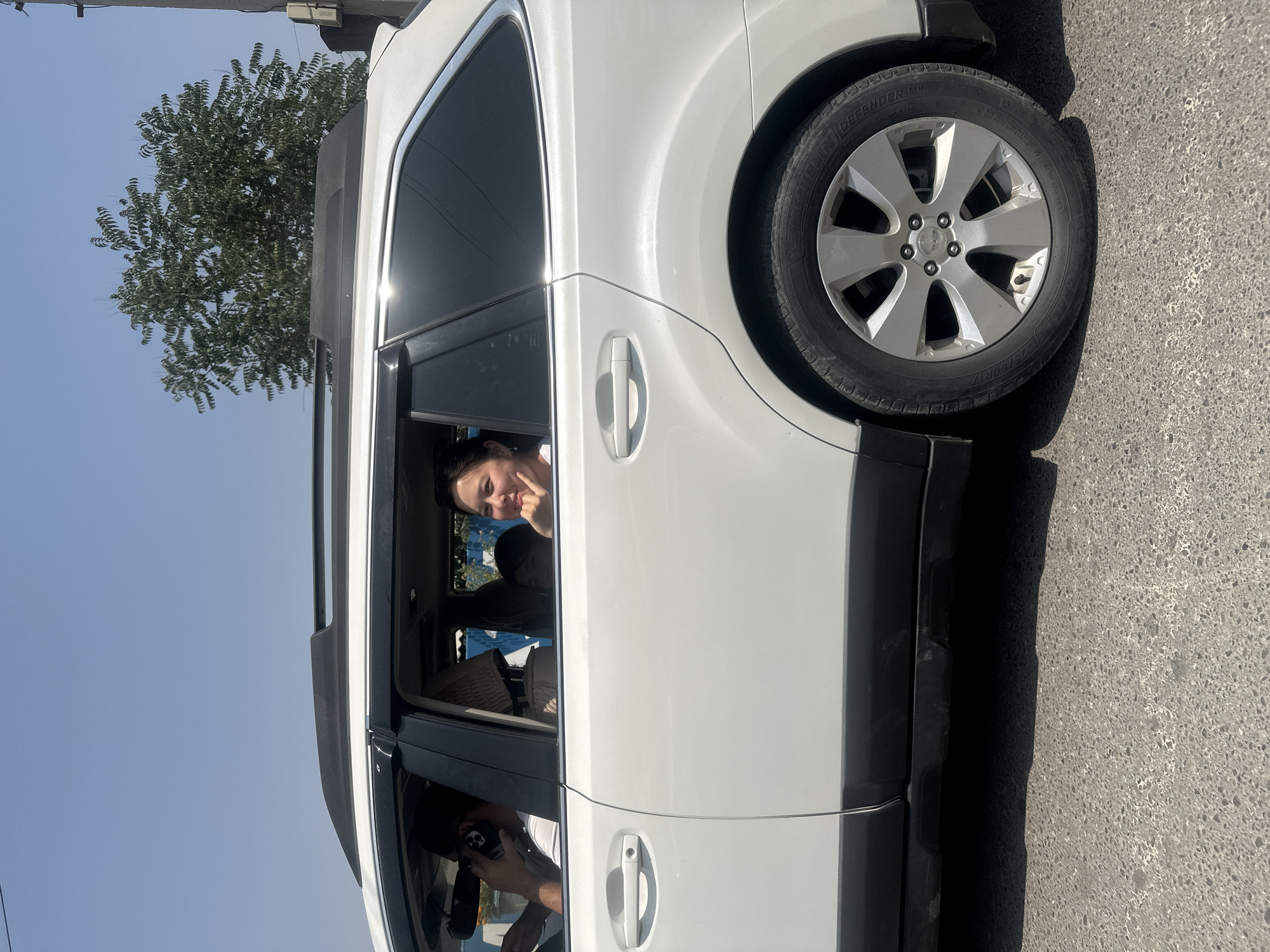 The image shows a white SUV parked on a paved road. Inside the car, a young woman with dark hair is visible through the open window, looking at the camera with a smile and her hand near her face. She appears to be in the passenger seat of the back row. Below her, in the front row, another person's head and shoulders are visible, holding a phone and taking a picture or video. The scene is set outdoors during the day, with a clear blue sky. A tree with green foliage is visible to the left of the vehicle, and the car's tire and part of its body are in focus on the right. The lighting suggests it is a sunny day, possibly late morning or early afternoon. The location appears to be a street in Manas, Kyrgyzstan, as indicated by the context. The overall mood seems lighthearted and casual, with the woman in the back appearing playful.