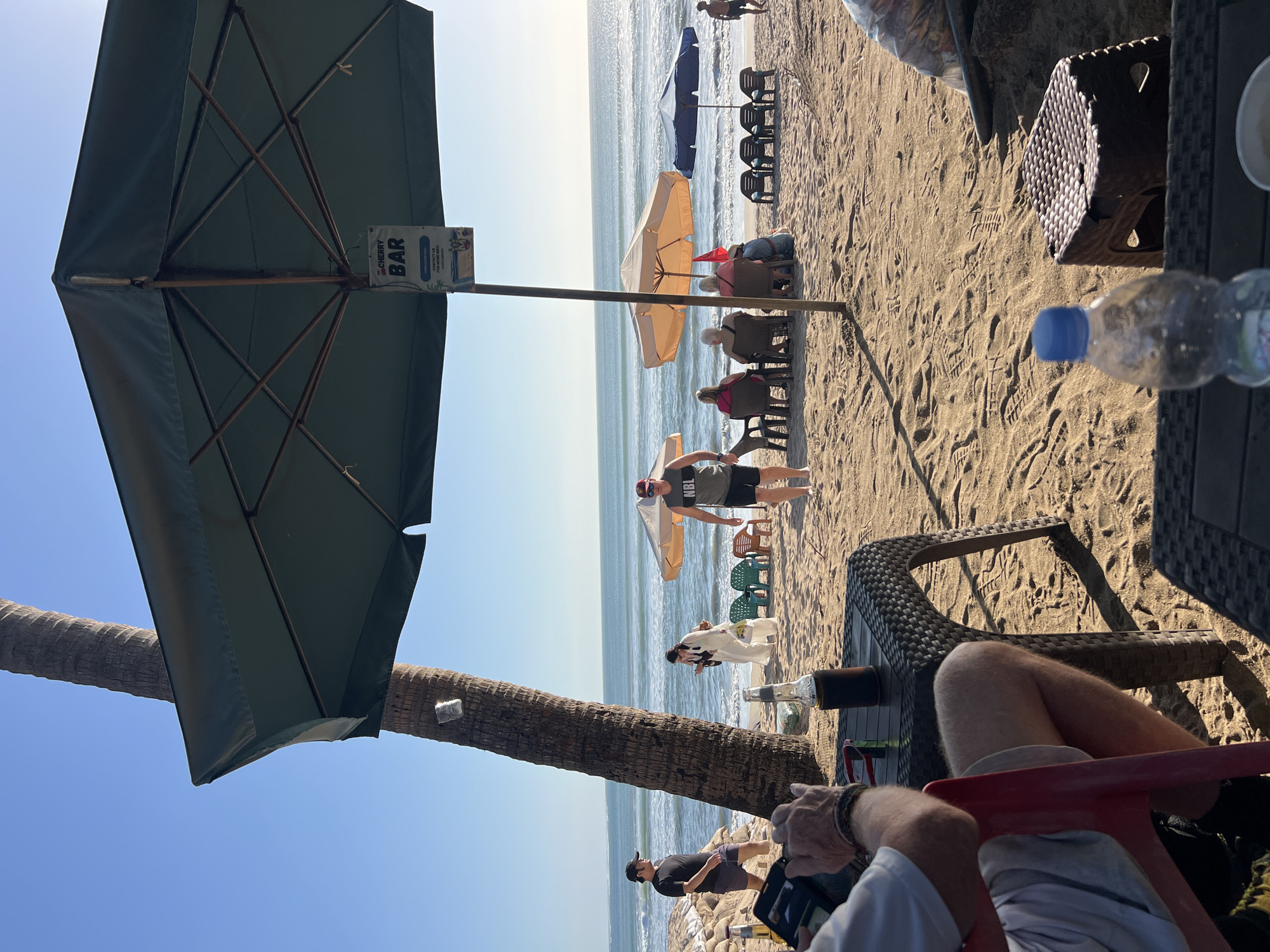The image captures a sunny day on Kuta Beach, Bali, Indonesia. In the foreground, a person is seated at a woven plastic table, holding a phone. On the table, there is a bottle of what appears to be dark liquid. To the right, a plastic stool and a plastic water bottle are visible. The sandy beach stretches towards the ocean, with waves gently rolling in. Several beach umbrellas are set up, one large green one dominating the upper portion of the frame, with a sign that reads "CHERRY BAR OPEN EVERYDAY CONTACT US FOR MORE INFO". Beneath this umbrella, a man in a gray tank top and black shorts is walking away from the viewer. Further back, under a smaller orange umbrella, a group of people are seated on chairs. Another blue umbrella is visible to the right, with several empty chairs in front of it. A palm tree stands tall on the left side of the image, leaning towards the sea. The sky is clear and blue. People are casually dressed, indicating a relaxed beach atmosphere.