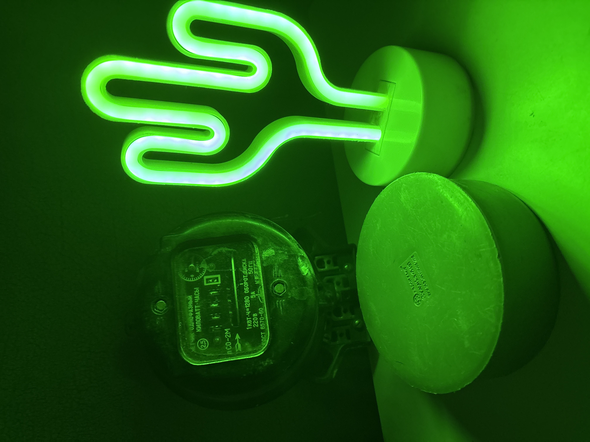 An overhead shot captures a dark, green-lit scene where a vintage kilowatt-hour meter sits to the left of a bright green neon cactus lamp. The meter, a dark, circular object, displays Russian text and has a circular dial with numbers. To its left, a round, dark object with embossed text sits on the surface. The neon cactus lamp, shaped like a stylized saguaro, emits a vibrant green light and stands on a circular base. The overall environment is dimly lit with an intense green hue, suggesting artificial lighting. The background is a dark, blurred green. The scene is indoors, likely on a flat surface such as a table or shelf.