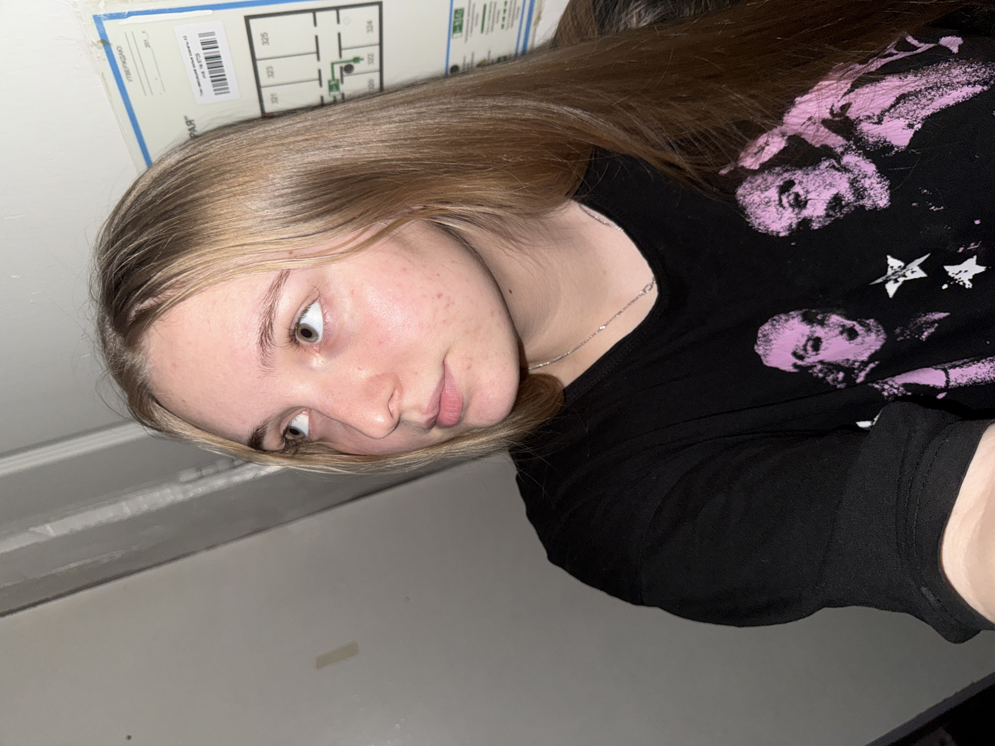 A young woman with long, blonde hair, wearing a black t-shirt with a pink graphic print, looks to her left. Her head is turned, and her eyes are directed upward and to the left. The background consists of a light-colored wall and a portion of a blue and white document that appears to be a floor plan or map, with the text "УТВЕРЖДАЮ" (APPROVED) visible. The overall lighting is dim, suggesting an indoor setting. No specific location within the city of Perm, Russia, can be identified from the image.