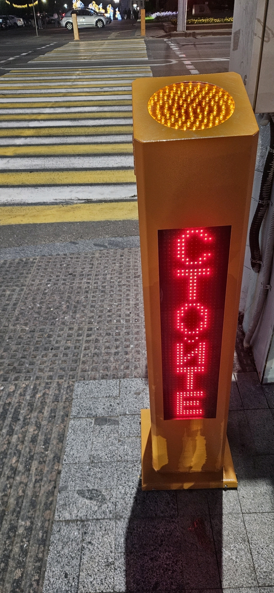 The image depicts a pedestrian crossing signal located in the city of Vitsebsk, Belarus. The signal is a tall, yellow, rectangular post with a red LED display. The display currently shows the word "СТОЙ" (which translates to "STOP" in Russian), indicating that pedestrians should not cross. Above the LED display, there is a circular amber light, likely a flashing warning signal. The pedestrian crossing itself is marked with prominent yellow and white stripes on the asphalt. In the background, a silver car is visible on the road, and streetlights illuminate the scene, suggesting it is nighttime or dusk. The ground around the signal is paved with textured grey tiles.