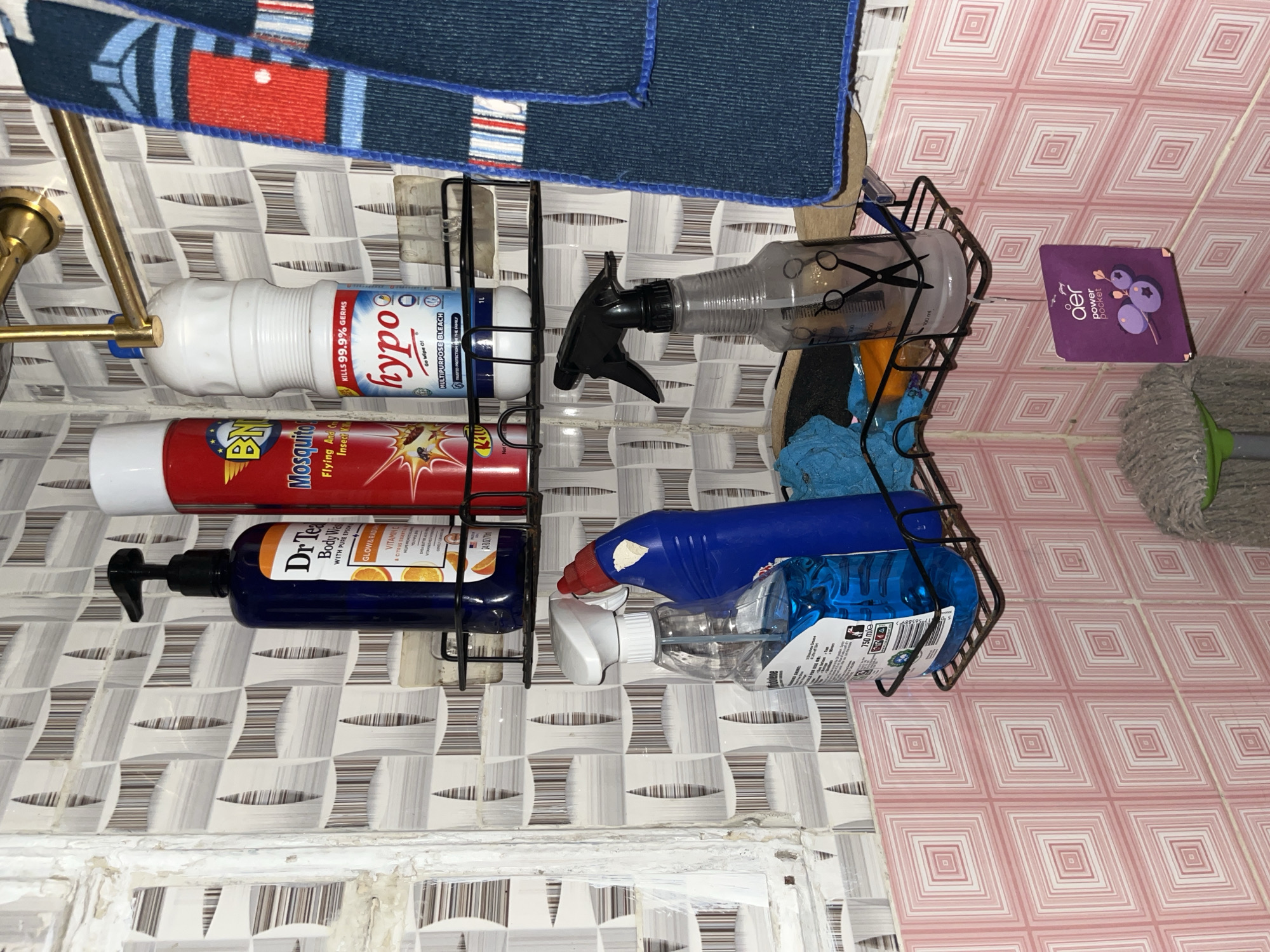 The image depicts a bathroom shelf in Abuja, Nigeria, filled with cleaning supplies and toiletries. On the top shelf, there are three bottles: a dark blue bottle of "Dr. Teal's Body Wash," a red can labeled "Mosquito Flying And Other Insect Killer," and a white bottle of "hypo MULTIPURPOSE BLEACH." To the right of these bottles, a blue towel with red and white stripes is hanging from a gold rod.
The bottom shelf holds two spray bottles: a clear bottle with blue liquid and a white spray nozzle, and a black spray bottle with a clear, partially filled reservoir that has a scissor graphic on it. Behind these bottles, a blue cleaning cloth is visible. Below the shelf, a small purple packet labeled "aer power pocket" with blueberry graphics is hanging. A mop head with a green handle is visible at the bottom of the image. The walls are tiled with a patterned design of white and grey geometric shapes. The overall scene suggests a domestic bathroom setting where cleaning products are stored.