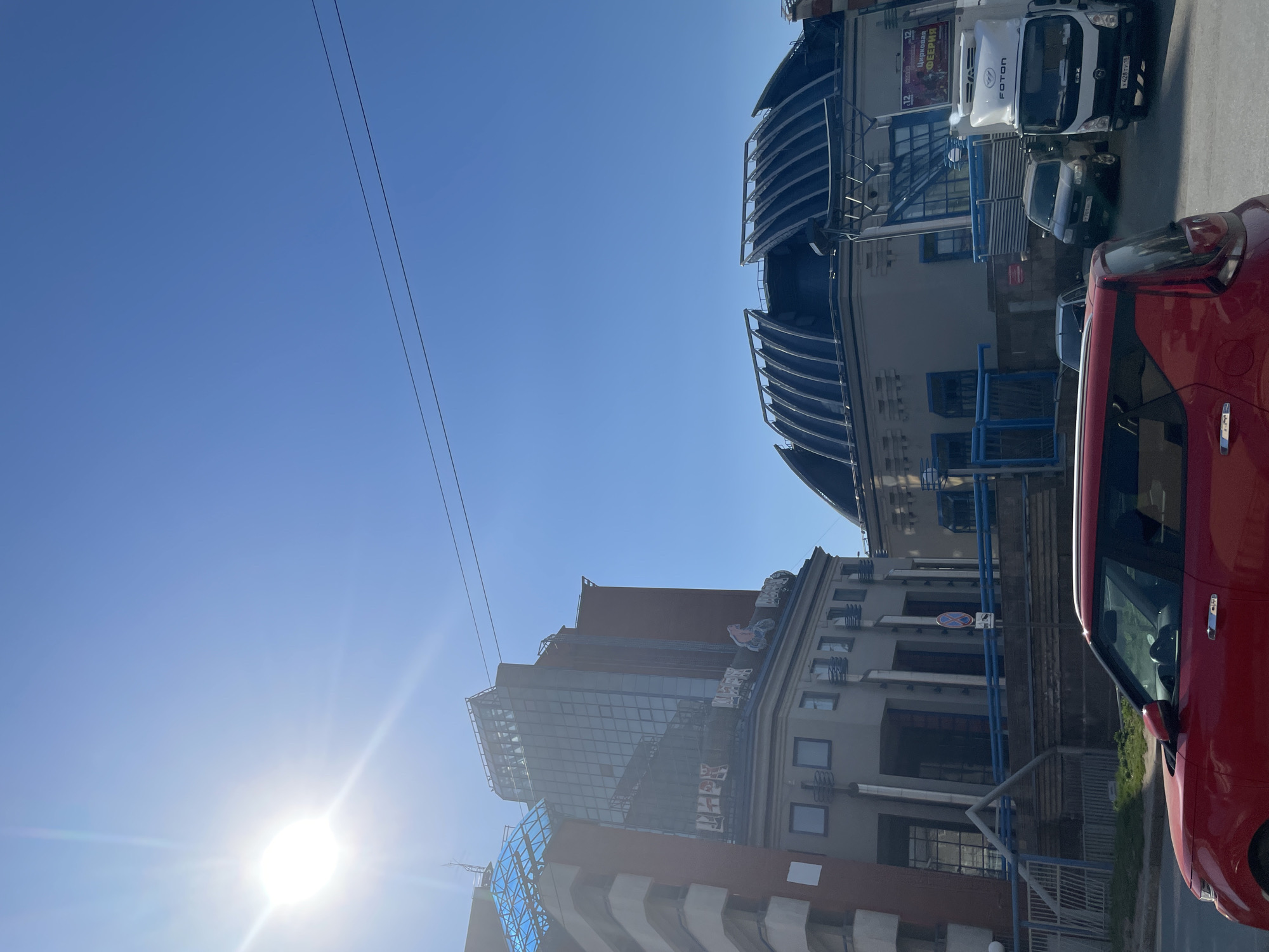 The image shows a street scene in Izhevsk, Russia, with buildings visible. On the right side of the image is a red car, and behind it, a white truck is parked. A smaller dark-colored car is parked near the truck. The buildings are multi-storied with architectural details like columns and decorative roofs. A blue sky with power lines stretches above. The sun is shining brightly in the lower-left corner of the image.
