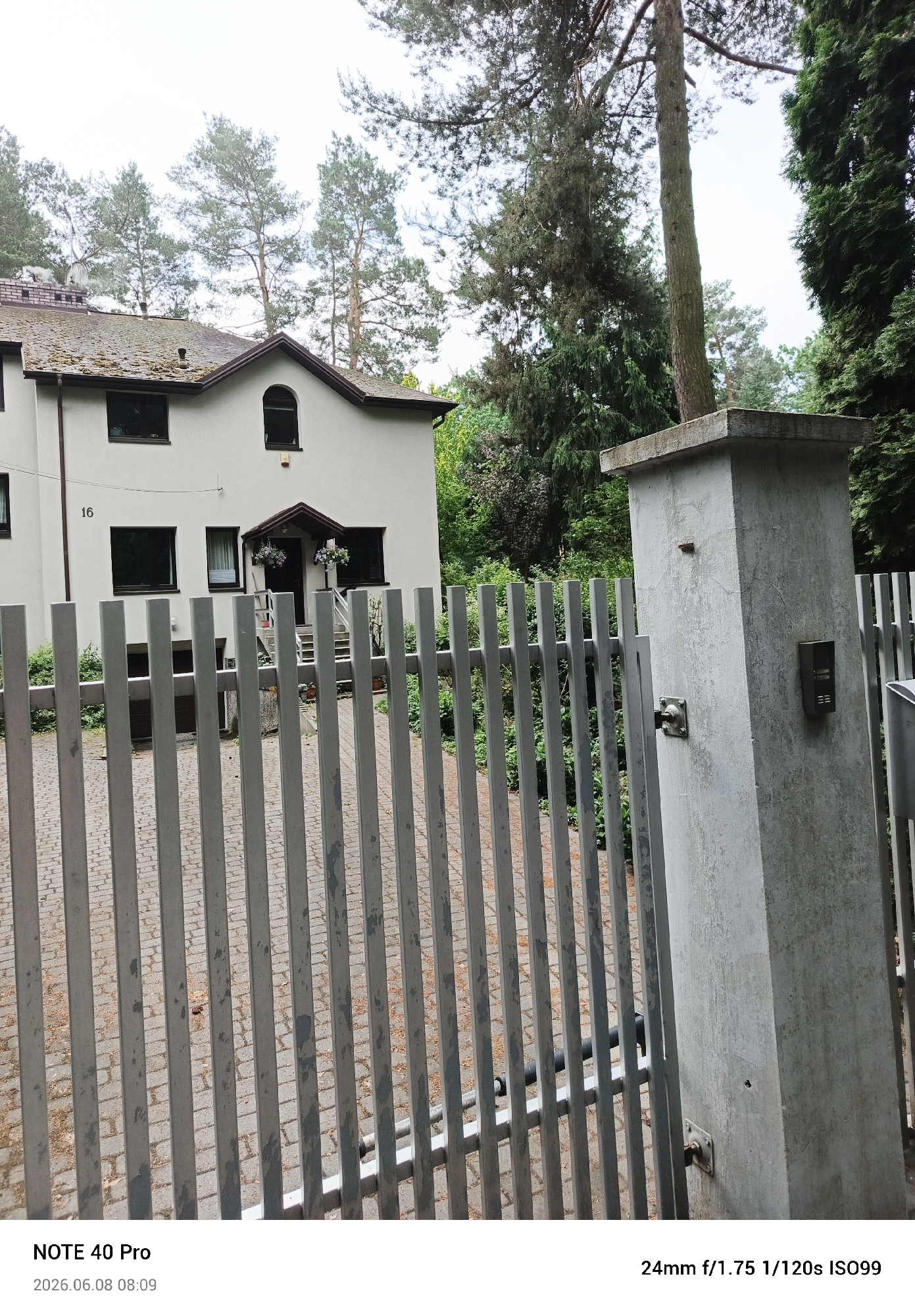 The image shows a view of a house with a fence in the foreground. The house is light-colored with dark-framed windows and a dark-colored roof. A number "16" is visible on the left side of the house. The fence is made of vertical metal bars. To the right of the fence, there is a concrete pillar with an intercom system and a door handle. The environment appears to be outdoors, with trees visible behind the house and to the right. The ground in front of the fence is paved with bricks. The lower portion of the image displays text identifying the camera as "NOTE 40 Pro" and the date and time as "2026.06.08 08:09", along with camera settings "24mm f/1.75 1/120s ISO99". The scene is consistent with a residential property.