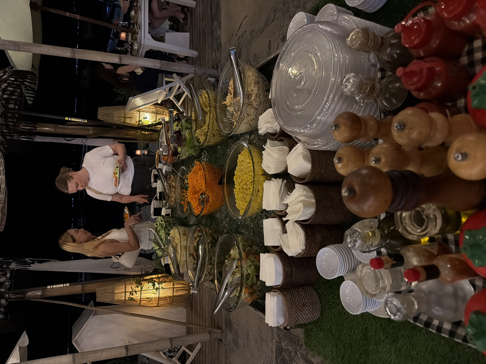 This image captures a buffet spread set up outdoors at night, likely in a tropical location given the bamboo structures and soft lighting. The buffet table is laden with various dishes, including salads in glass bowls filled with ice. Visible salads include shredded carrots, corn, pasta, and leafy greens. Several servers are present, with two women at the front of the buffet selecting food. One woman, wearing a white top and a white purse, points to a dish. The other woman, in a white t-shirt and dark pants, holds a plate. In the background, people are seated at tables, suggesting a dining area. The environment appears to be a beachfront or waterside setting, with water and lights visible in the distance. The overall scene suggests an evening event with a self-service food offering.