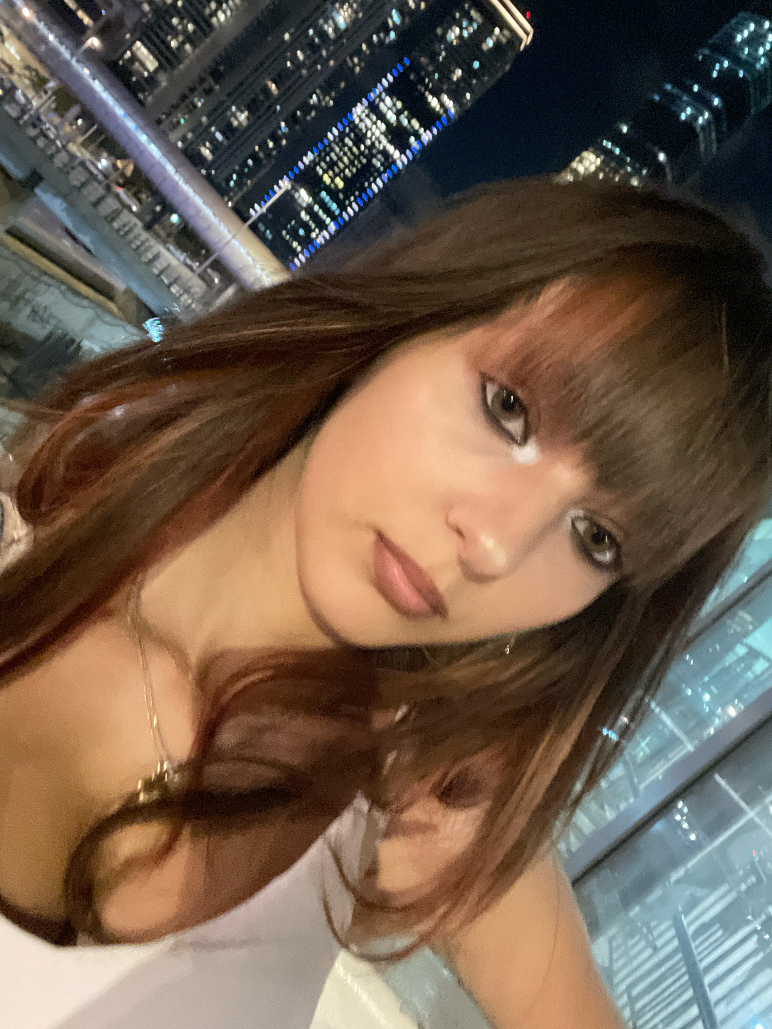 A young woman with long brown hair and bangs is shown in a selfie taken at night. She is wearing a necklace. In the background, city lights from high-rise buildings are visible. The scene appears to be outdoors, possibly on a balcony or rooftop, overlooking a cityscape. It is not possible to determine the specific location within Tel-Aviv, Israel from the image alone.