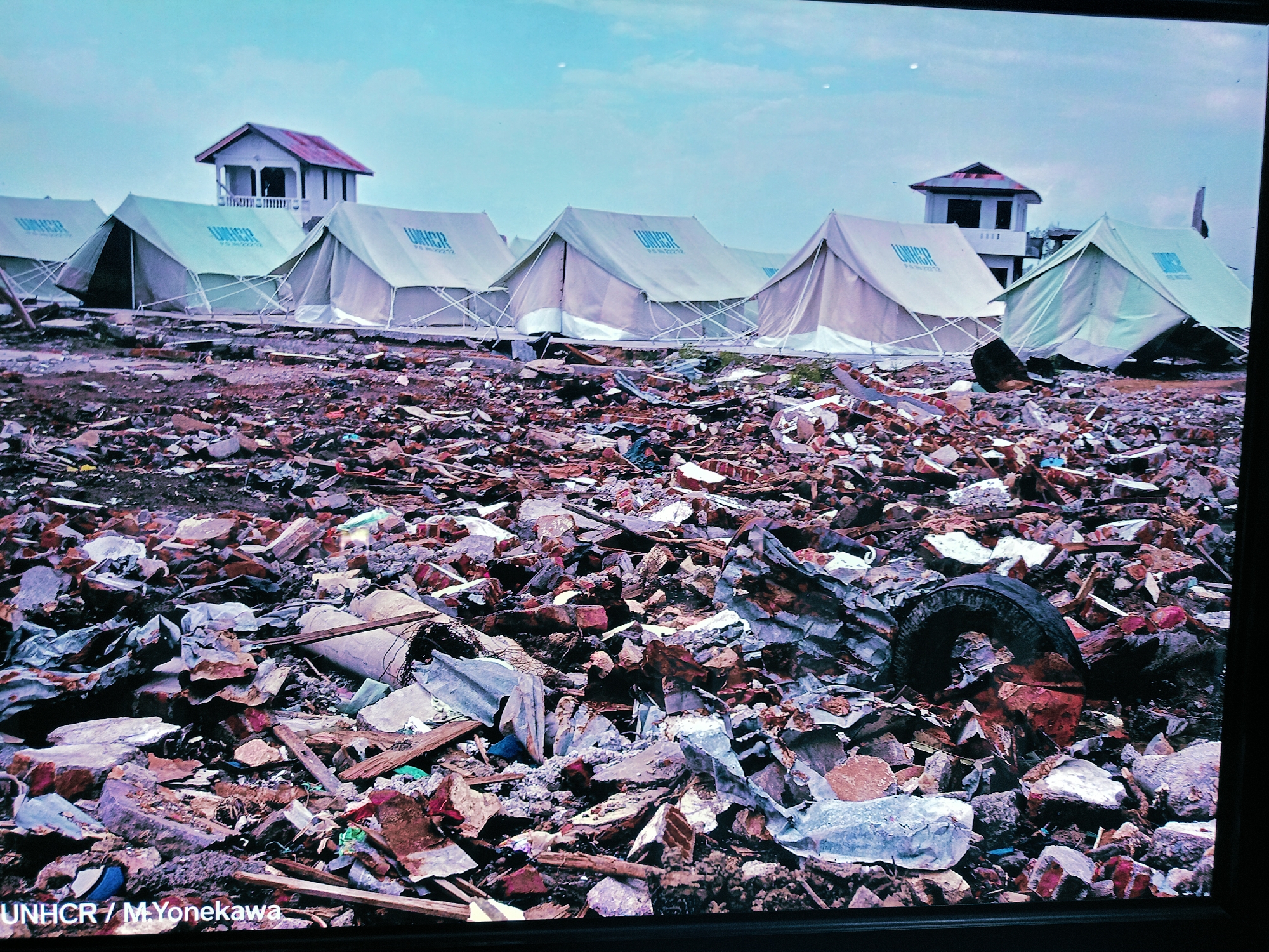 The image depicts a scene of devastation, likely in Banda Aceh, Indonesia, after a natural disaster. The foreground is covered in a chaotic jumble of debris: broken bricks, concrete fragments, twisted metal, and various unidentified objects, including a tire. In the background, a row of white UNHCR (United Nations High Commissioner for Refugees) tents stand on cleared ground. Behind the tents, two small buildings or guard towers remain standing, appearing relatively intact. The sky is overcast. No people are visible. The scene conveys a sense of large-scale destruction and the subsequent provision of emergency shelter.