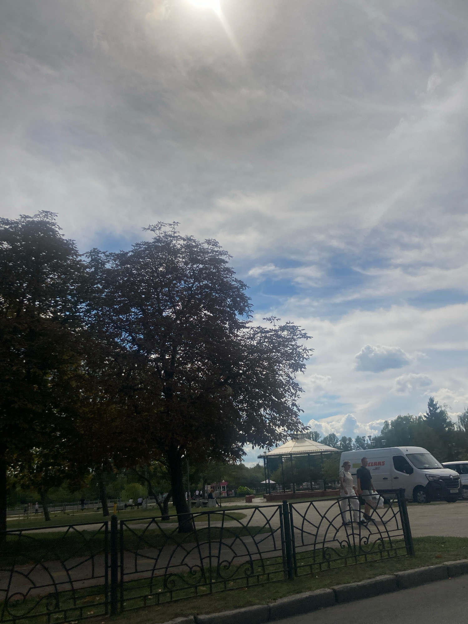 The image depicts an outdoor scene with a bright, overcast sky and sunlight filtering through thin clouds. In the foreground, a black metal fence with an ornate design runs horizontally across the frame. Behind the fence is a paved area, likely a road or parking lot, and a grassy verge.
Further into the background, several trees with dark foliage are visible. To the right, a white van with "CLAAS" branding is parked. Two individuals, a woman in a striped top and white pants and a man in a dark t-shirt and shorts, are walking together near the van.
In the middle ground, there is a park-like setting with more trees, benches, and a gazebo-like structure. Several other people are scattered throughout this area, some walking and some sitting. Further back, a playground with colorful structures can be seen. The overall environment suggests a public park or recreational area. The presence of the "CLAAS" branded van suggests that this location might be a commercial area or an event venue that uses such services. While no specific landmarks clearly identify Brest, Belarus, the overall scene is consistent with urban parkland found in many cities.