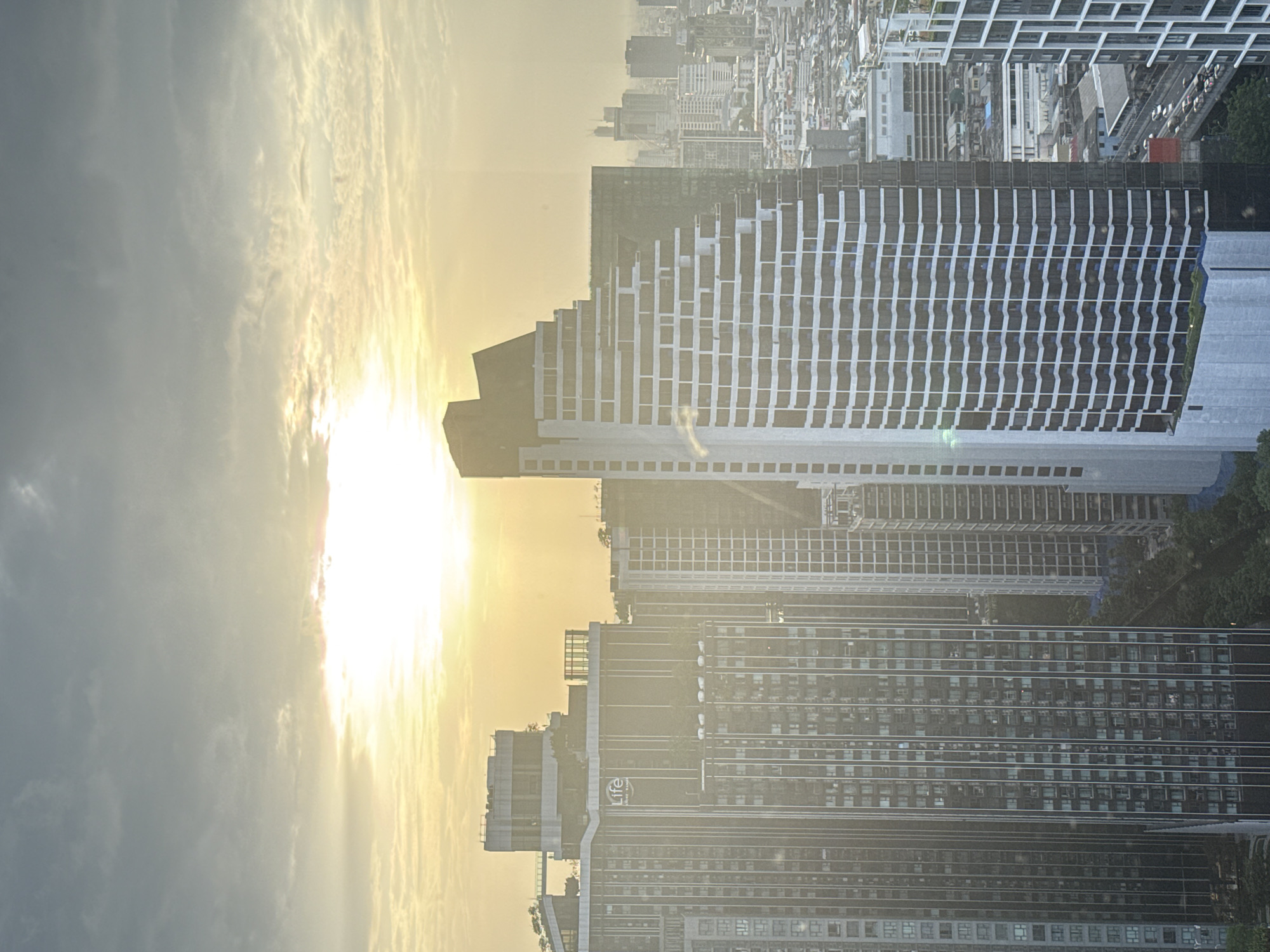 An aerial view captures a hazy sunset over Bangkok, Thailand, casting a warm, yellow glow across the cityscape. The sun, a bright, diffused orb, is partially obscured by clouds and atmospheric haze. In the foreground, a cluster of modern skyscrapers dominates the view. A distinctive, uniquely shaped building with a stepped, triangular facade stands prominently on the right, its white balconies creating a textured pattern. To its left, a taller, darker building with a grid-like pattern of windows and vertical white accents is visible. Further left, another imposing skyscraper with vertical white lines and numerous windows is seen. The middle ground reveals a denser collection of buildings, some with dark facades and others with white, giving a sense of depth and urban sprawl. In the background, more buildings, some appearing lighter and more spread out, fade into the hazy distance, suggesting the vastness of the city. The overall impression is a dense urban environment bathed in the soft light of sunset.