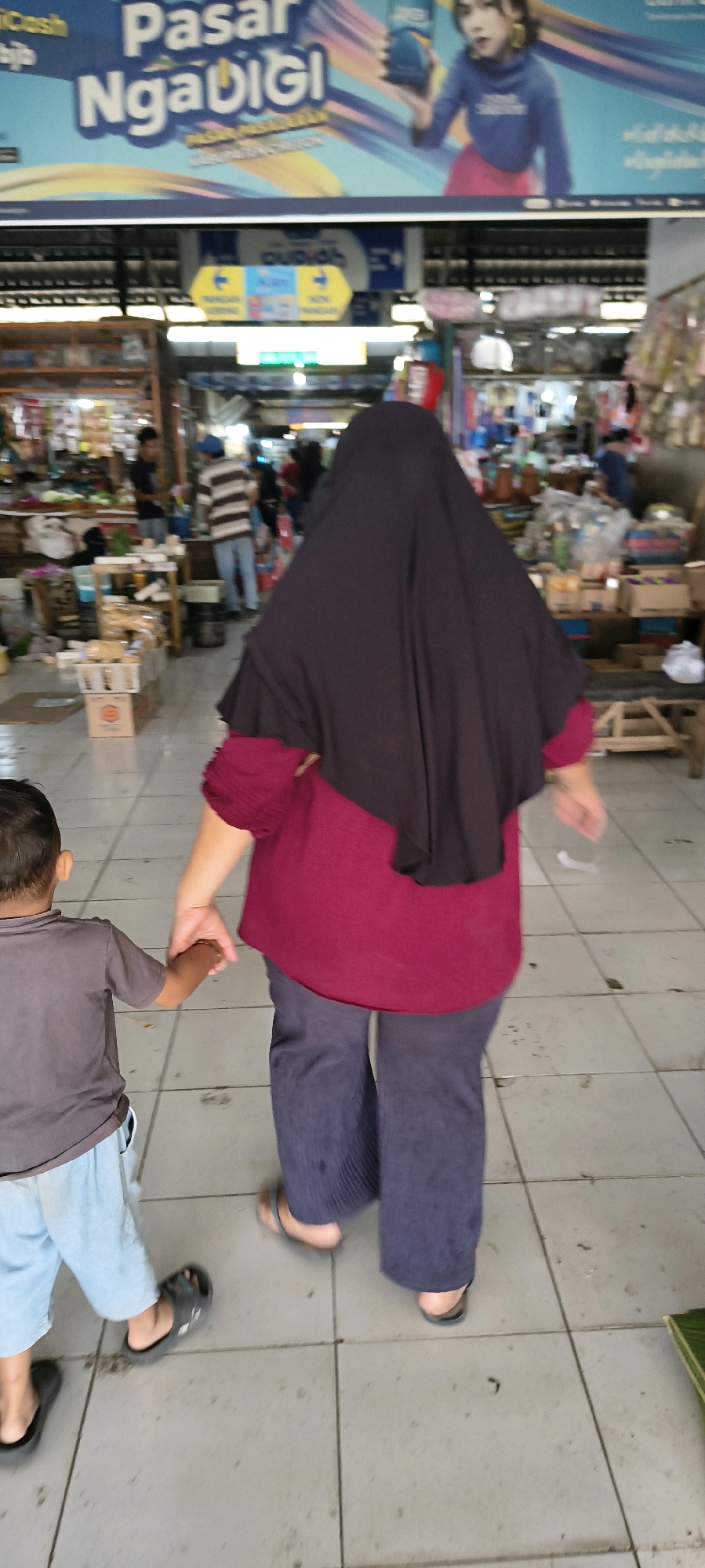 In the city of Weru Lor, Indonesia, a person wearing a dark hijab and a maroon shirt is walking with a young child, holding their hand. The child is wearing a grey t-shirt and light blue shorts. They are walking on a tiled floor inside a marketplace. In the background, stalls are visible, with goods displayed on shelves and tables. A sign with Indonesian text, including "Pasar Ngadigisi," is visible at the top of the frame, indicating the location as a market. Other people can be seen in the background, engaged in various activities within the market. The overall scene depicts a casual moment within a bustling Indonesian market.