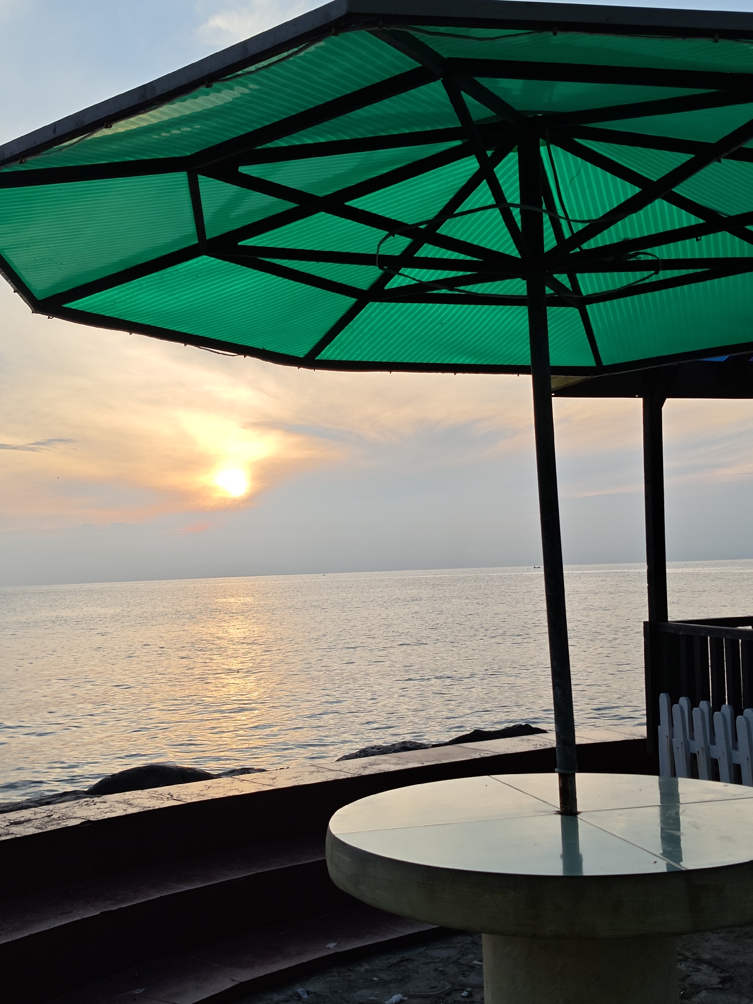A wide shot captures a tranquil beach scene at sunset in Lhokseumawe, Indonesia. In the foreground, a round, light-colored table with a reflective surface stands on sand. Behind it, a large, green umbrella with a metal frame provides shade, supported by a single dark pole. The umbrella's corrugated surface is visible, along with its internal support structure. A low, dark-colored wall runs along the edge of the beach, with a few white posts and a dark wooden fence visible further back.
The middle ground is dominated by the calm sea, its surface reflecting the soft hues of the setting sun. The sun itself is a bright, diffused orb in the sky, casting a warm glow across the water and clouds. The horizon line is distinct, separating the sea from the pale sky, which is streaked with subtle shades of orange and pink from the sunset. The overall atmosphere is peaceful and serene.