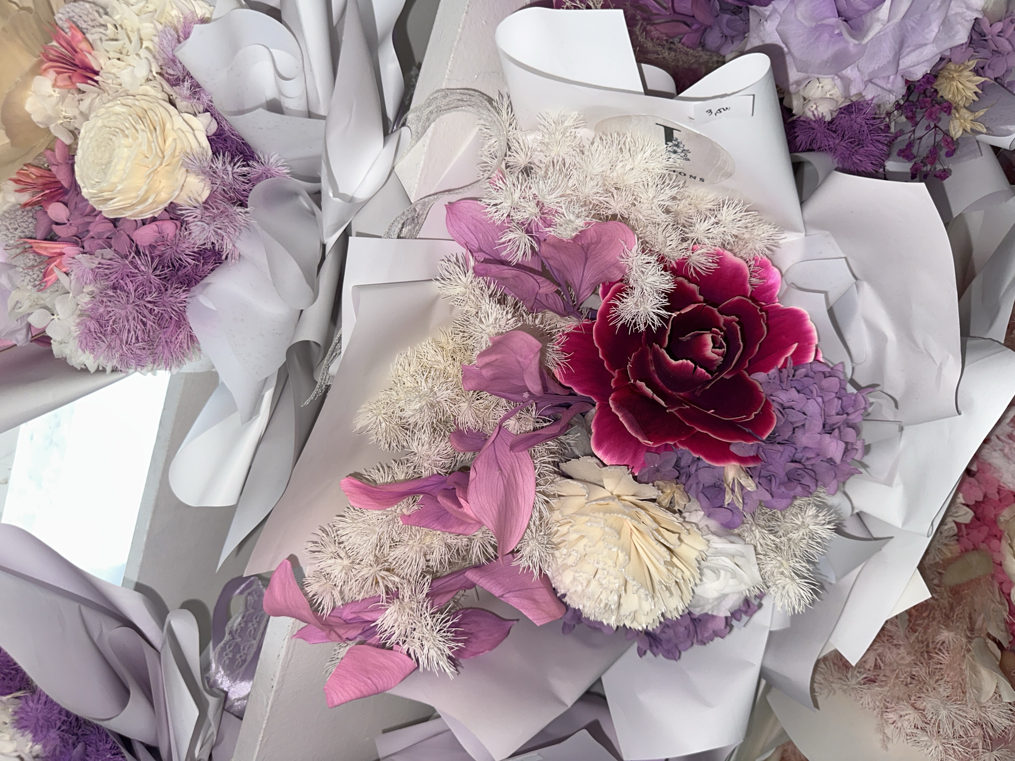The image displays a close-up view of several bouquets of dried and preserved flowers. The bouquets are arranged in a way that suggests they are either for sale or on display. The dominant colors are shades of purple, pink, cream, and deep red, with accents of white and light green.
In the foreground, a prominent bouquet features a large, deep red flower with ruffled petals at its center. Surrounding this flower are delicate, spiky white elements, soft pink leaf-like structures, and clusters of small purple flowers. To the left and slightly behind this central bouquet, another bouquet is partially visible, showcasing similar white spiky elements and purple floral clusters, along with a large, textured cream-colored bloom. In the upper right of the image, a portion of a third bouquet is visible, also featuring purple and cream-colored preserved flowers. The bouquets are wrapped in white paper, with some sections of lavender-colored paper peeking through.
The background is out of focus but appears to be a brightly lit, possibly outdoor or windowed area. The lighting suggests daylight, with some blown-out highlights indicating a strong light source. There are no people visible in the image, and no specific geographical markers or elements in the scene can definitively place it in Davao City, Philippines, or any other specific city. The focus is entirely on the floral arrangements.