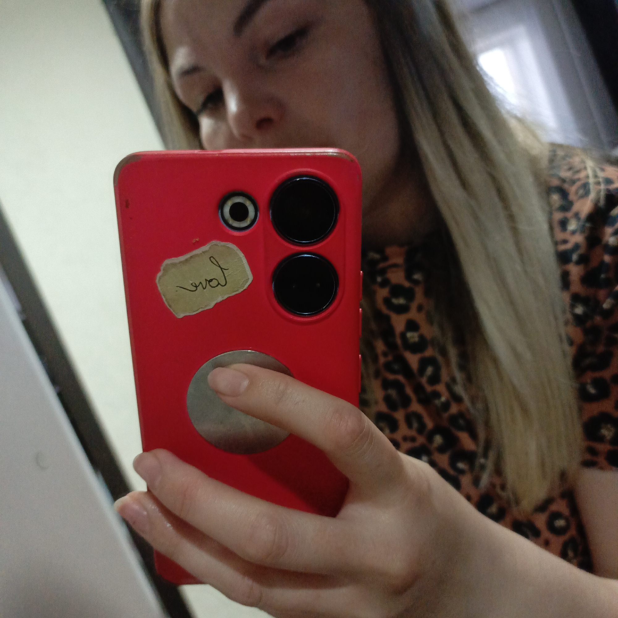 A woman is depicted taking a mirror selfie with a bright red smartphone. Her face is partially visible in the reflection, showing her looking downwards towards the phone. She has long, blonde hair and is wearing a brown and black leopard print top.
Her right hand holds the phone, with her thumb resting on a circular, metallic attachment (possibly a pop socket or magnetic mount) near the bottom of the phone case. The red phone case features two prominent black circular camera lenses stacked vertically, with a smaller circular camera or flash unit positioned above them. A small, light-colored sticker with torn edges is adhered to the case, displaying the word "eval" in black cursive script, which is "love" mirrored due to the reflection.
The background is blurry, indicating an indoor setting reflected in the mirror. Vague shapes and light patches suggest a room interior, possibly including a window or light source in the upper right of the frame. The image has a slightly grainy quality.