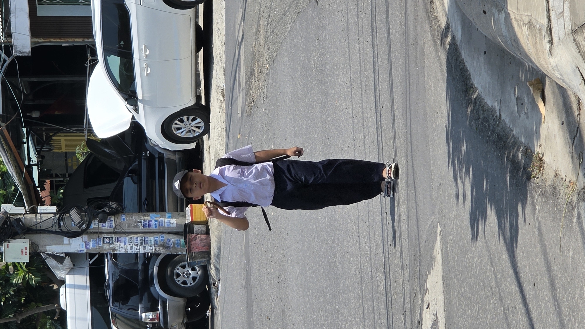 A medium, outdoor shot shows a person wearing a short-sleeved white shirt, dark pants, and a dark baseball cap, standing in the middle of a gray asphalt street in Bac Nha Trang Ward, Vietnam. The person is facing forward with their right arm raised, holding a brown object, and their left arm at their side. The person appears to be in their youth, and is wearing a black backpack. The street is bordered on the left by parked vehicles, a utility pole plastered with paper, and vegetation. The vehicles include a white minivan, a black SUV, and a white truck. On the right, the street is bordered by a concrete retaining wall, from which a small amount of vegetation grows. The image is oriented vertically, but the scene appears to be tilted counter-clockwise.