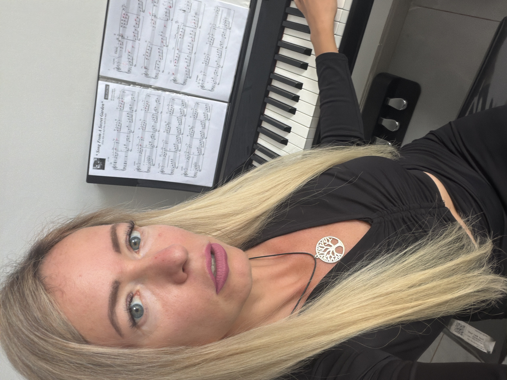 The image shows a blonde woman sitting at a digital piano. She is wearing a black ruched top and a black skirt. A silver tree of life pendant is visible on a black cord around her neck. Sheet music titled "Song From A Secret Garden" is displayed on the piano. The setting appears to be indoors, with a plain white wall in the background. There is no visible indication of the location being in Xuân Trường - Đà Lạt, Vietnam.
