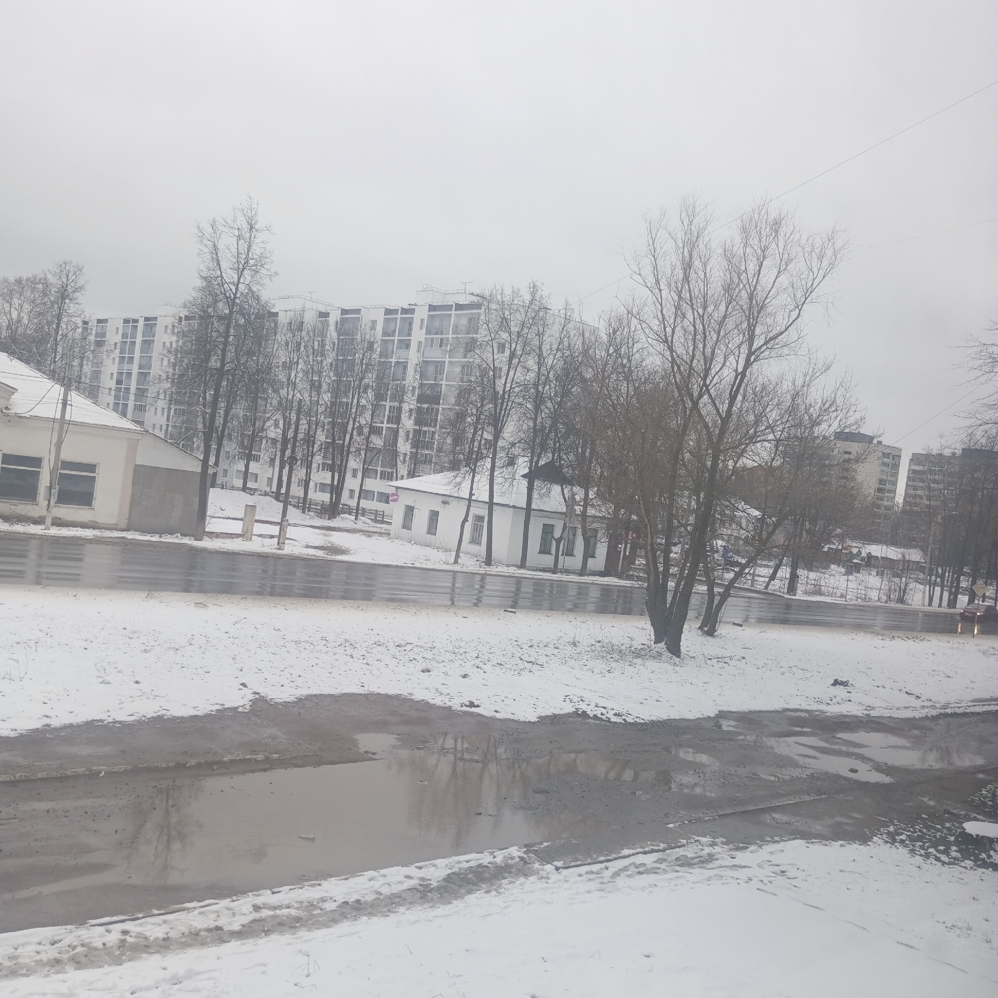 The image depicts a snow-covered street scene in Chaikovsky, Russia. The foreground features puddles of water reflecting the overcast sky. A low fence separates the foreground from the street. Buildings are visible in the midground, including a single-story structure and a taller apartment building in the background. Bare trees are scattered throughout the scene. A red car is visible in the distance to the right, but there are no people visible. The sky is uniformly gray, indicating a cloudy day.