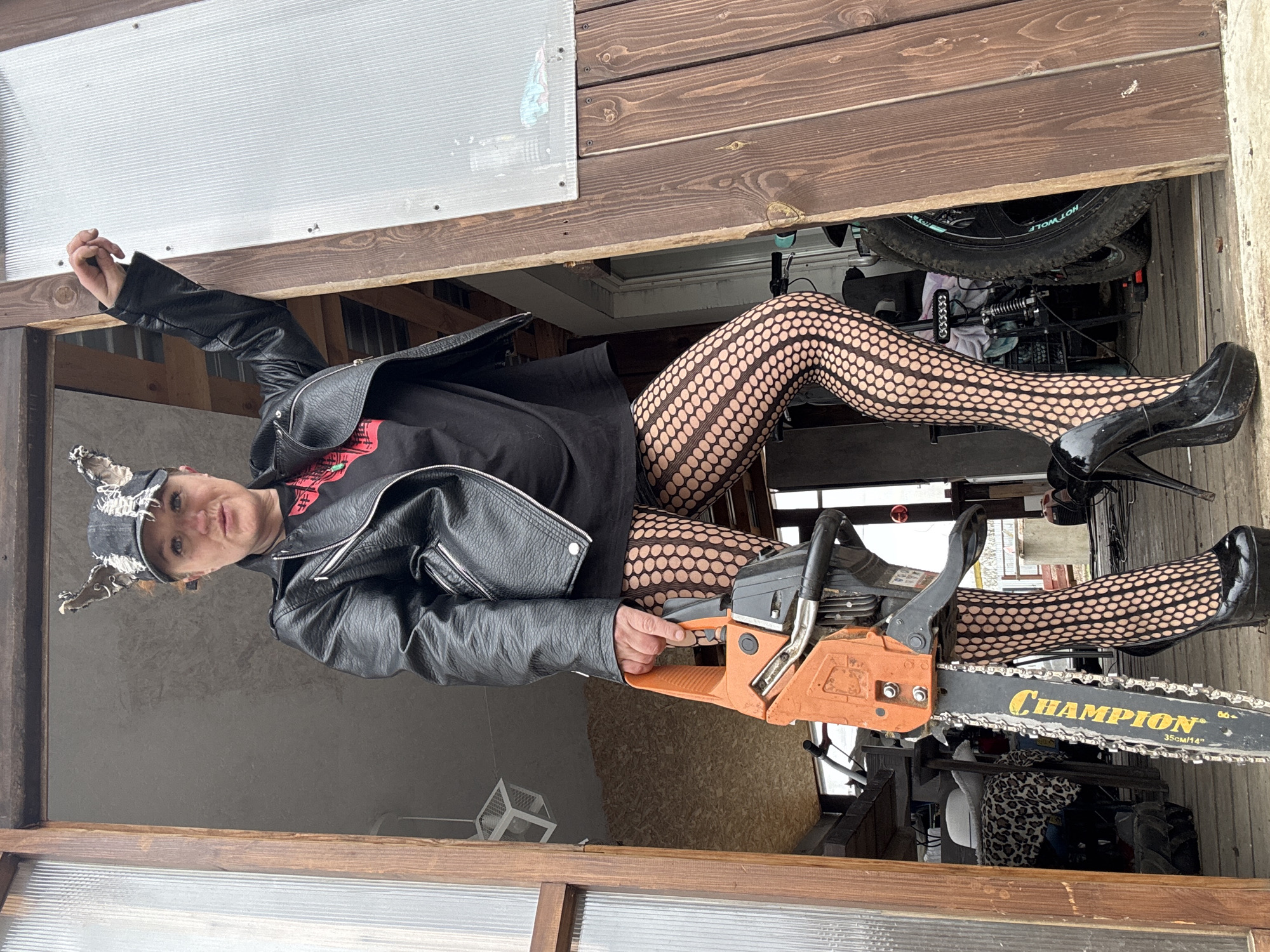 A person wearing a black leather jacket, a black shirt, black fishnet stockings, and black high heels is holding an orange and gray Champion chainsaw. The person is wearing a cap with decorative elements. The scene appears to be set in or near a garage or storage area, with exposed wooden beams and various items stored behind the person. Based on the camera angle, the person may be standing or sitting in an elevated position relative to the camera. The location is the city of Chaikovsky, Russia.