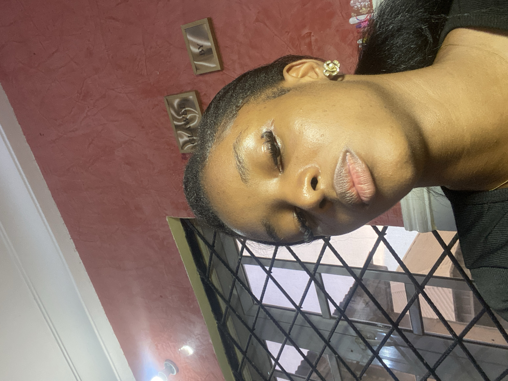 The image is a selfie taken by a young Black woman indoors, likely in Jos, Nigeria. The woman is looking down with her eyes closed and her lips slightly parted. She is wearing a black top and a gold earring in her right ear. The background is a red textured wall with two framed artworks hanging on it. To the left of the woman is a barred window, through which a building can be seen. A light fixture is visible on the ceiling above the woman. The overall lighting is dim, with a bright light source coming from the left side of the image, illuminating the woman's face.