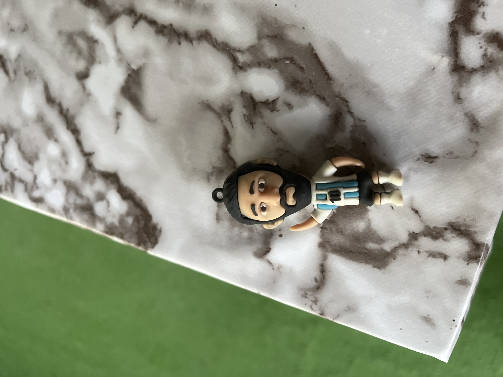 A small, detailed figurine of a soccer player, likely representing Lionel Messi, is positioned on a marble-patterned surface. The figurine is wearing an Argentina national team jersey with the number 10. Its right arm is raised in a waving gesture. The background consists of a blurred green surface. There are no discernible elements in the image that can be linked to the city of Vărzărești, Moldova.