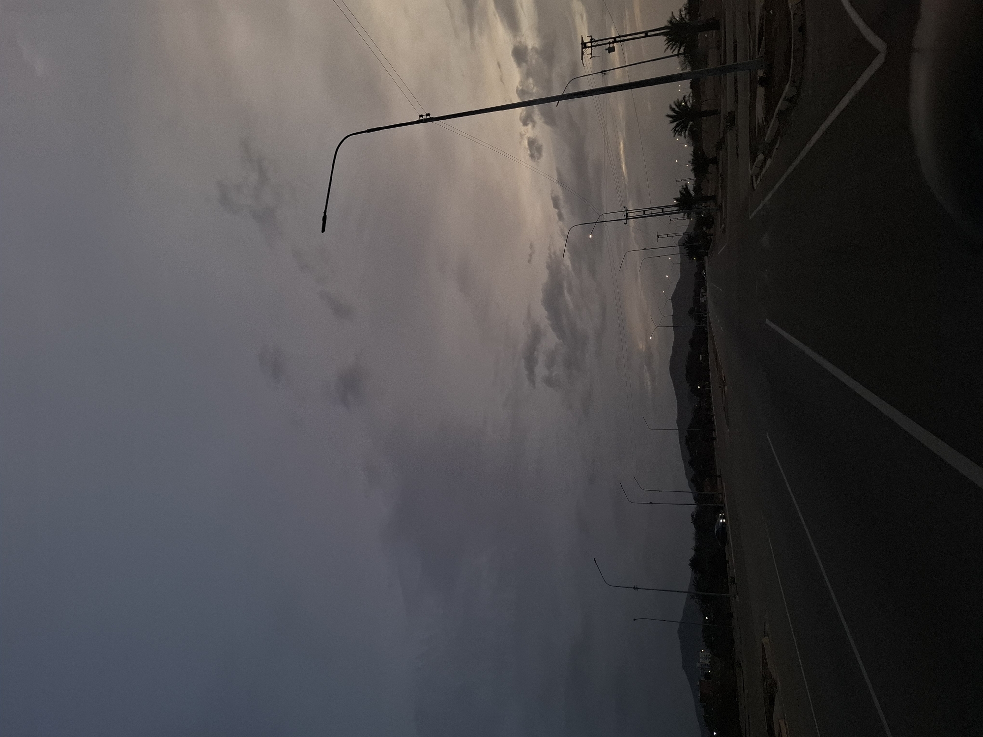 The image depicts an asphalt road with a single white lane divider. The road is flanked by streetlights and palm trees. The sky is overcast with dark, heavy clouds. In the background, low-lying mountains are visible. The image appears to be taken at dusk or dawn. The location is inferred to be Altagracia, Venezuela. There is a car on the right side of the road.
