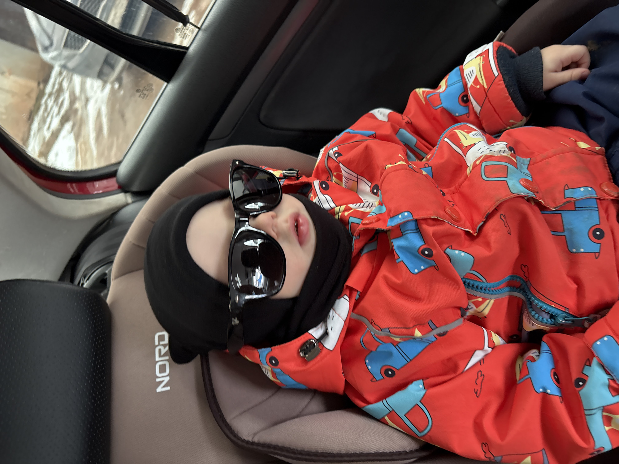 A toddler wearing a black balaclava and oversized sunglasses is lying in a light brown car seat with "NORD" printed in white letters on its side. The toddler is wearing a red and blue patterned jacket. The background shows the interior of a car, including the back window. The location is Chaikovsky, Russia.