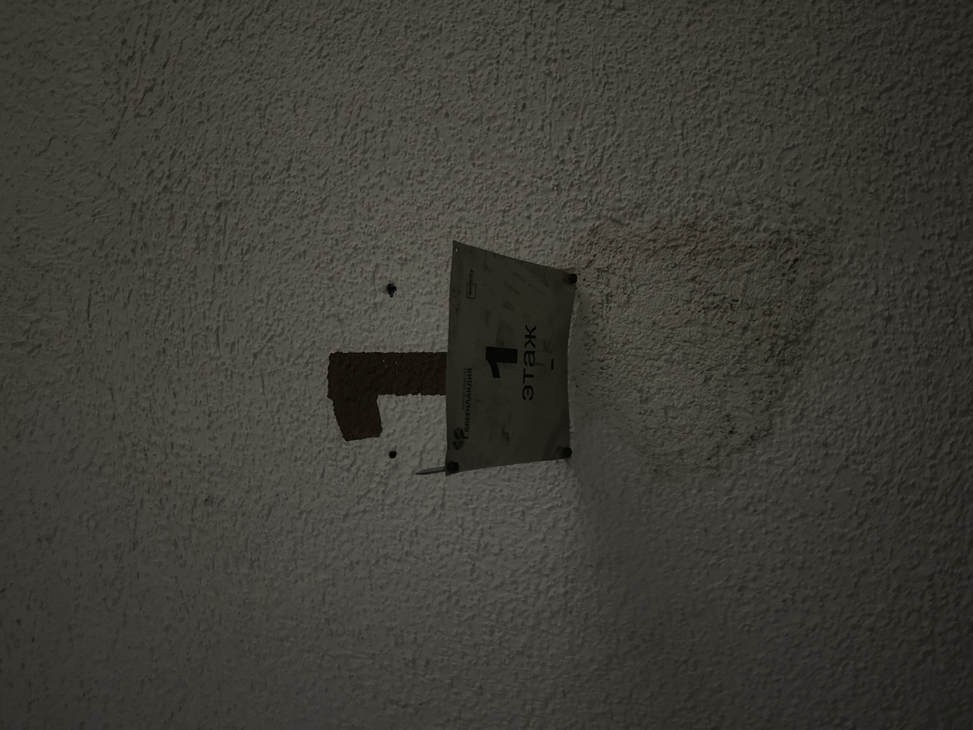 A close-up shot captures a textured, light-colored wall with a dark shadow cast on the lower right. Affixed to the wall is a sign with a large, dark brown number "1" painted above it. The sign itself is white and features text and a logo, appearing to be a building directory or floor marker. The text "GREENLANDIA" and "1" are visible, along with smaller Russian text that reads "этаж" (etazh), meaning "floor." The sign is attached to the wall with screws. The overall impression is that of an interior wall in a building. This image is located in the city of Sinyavino, Russia.
