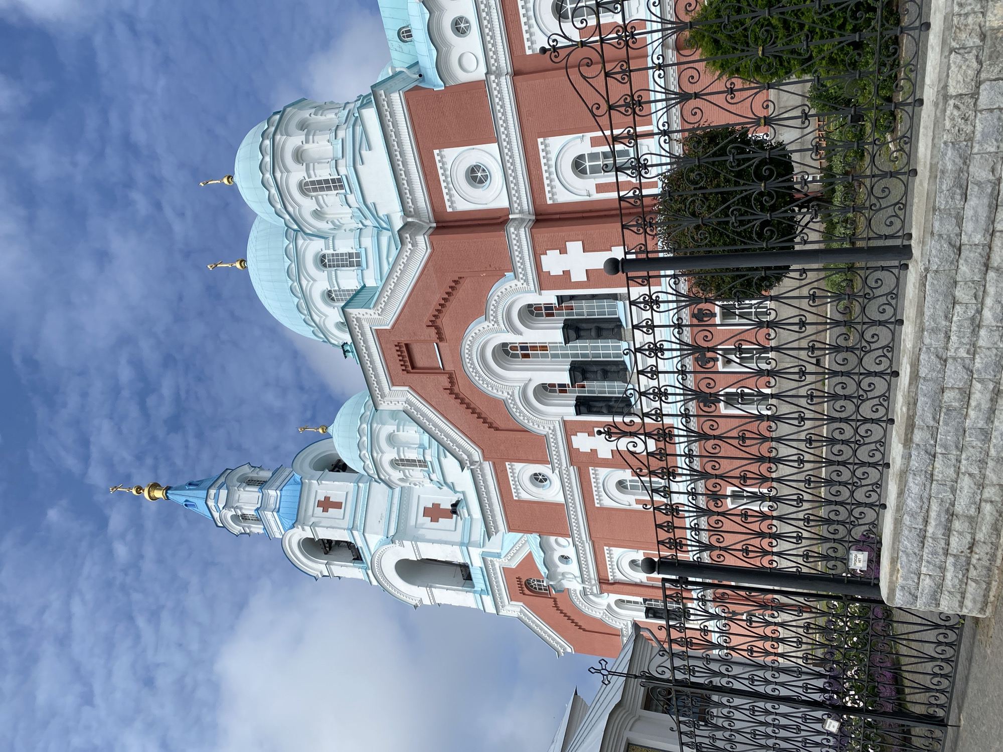 The image shows the exterior of a grand, pink and white Orthodox church with blue domes, located in Sортавала, Russia. A decorative black metal fence with a stone base stands in the foreground, partially obscuring the view of the church's lower sections. The church features multiple onion domes topped with golden crosses, a bell tower with arches and red crosses, and a facade adorned with white decorative elements and arched windows. The sky is blue with scattered white clouds. No people are visible in the image.