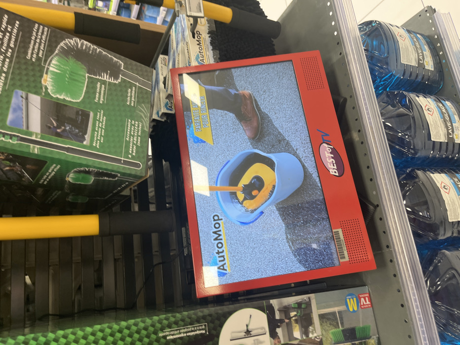 The image shows a red, small TV screen playing a commercial for an Auto Mop on a shelf in a retail store located in Saint-Brice-Courcelles, France (latitude: 49.283, longitude: 4). The TV screen shows a person's foot in brown shoes stepping on a gray, textured surface next to a blue bucket filled with soapy water and a yellow mop handle. The mop head is partially submerged in the water, and a yellow mop handle is visible. The TV screen is tilted forward and slightly to the right. The screen is on a black shelf with white dots. Above the TV screen are product boxes featuring pictures of cleaning tools and accessories. Three blue containers of glass cleaner are visible below the shelf. The container labels say "LAVE-GLACE". One container, partially visible, says "ACE".