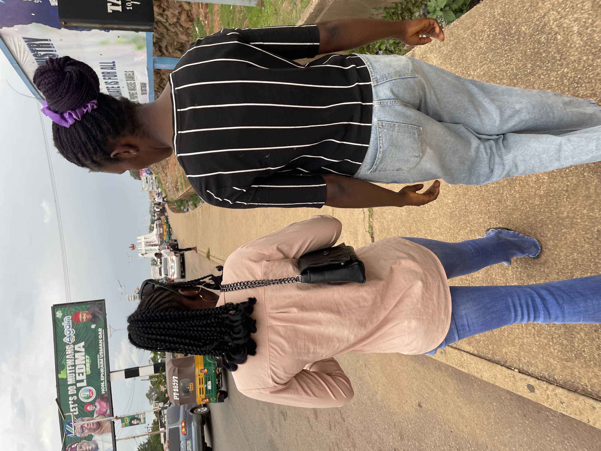 Two women walk away from the camera on a sidewalk in Jos, Nigeria. The woman on the left wears a light pink button-down shirt and blue jeans. She has braided hair and carries a black purse over her shoulder. The woman on the right wears a black and white striped t-shirt and light blue jeans. Her hair is braided and pulled into a bun with a purple scrunchie. They are walking past a busy street with cars, motorcycles, and billboards. One billboard features a campaign slogan in English and images of political figures. The sky is overcast.
