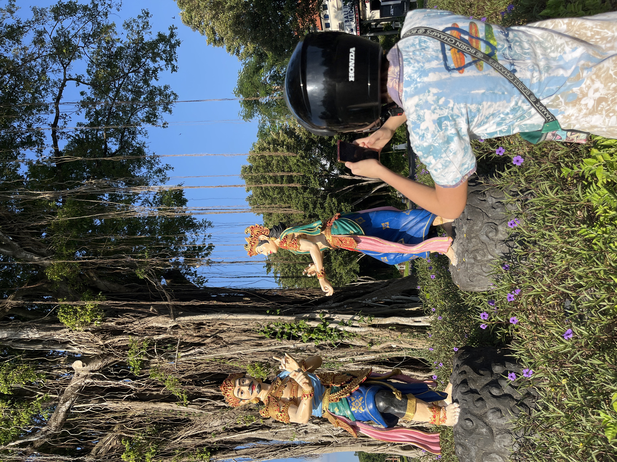 In Kuta Selatan, Indonesia, a person wearing a black Honda helmet and a tie-dye shirt is holding a smartphone to take a picture of two large, colorful statues. The statues depict a male and female figure in traditional Balinese attire, standing on dark, textured bases amidst lush green foliage and small purple flowers. Behind the statues, a massive banyan tree with a thick trunk and numerous aerial roots dominates the scene. The tree's branches extend upwards, covered in green leaves, and hang down in long tendrils, creating a dramatic backdrop. The sky above is a clear, bright blue. In the background, a building with signage that includes "BALI INT" and "MED" is visible, along with other trees and vegetation. The overall environment suggests an outdoor cultural or tourist attraction.