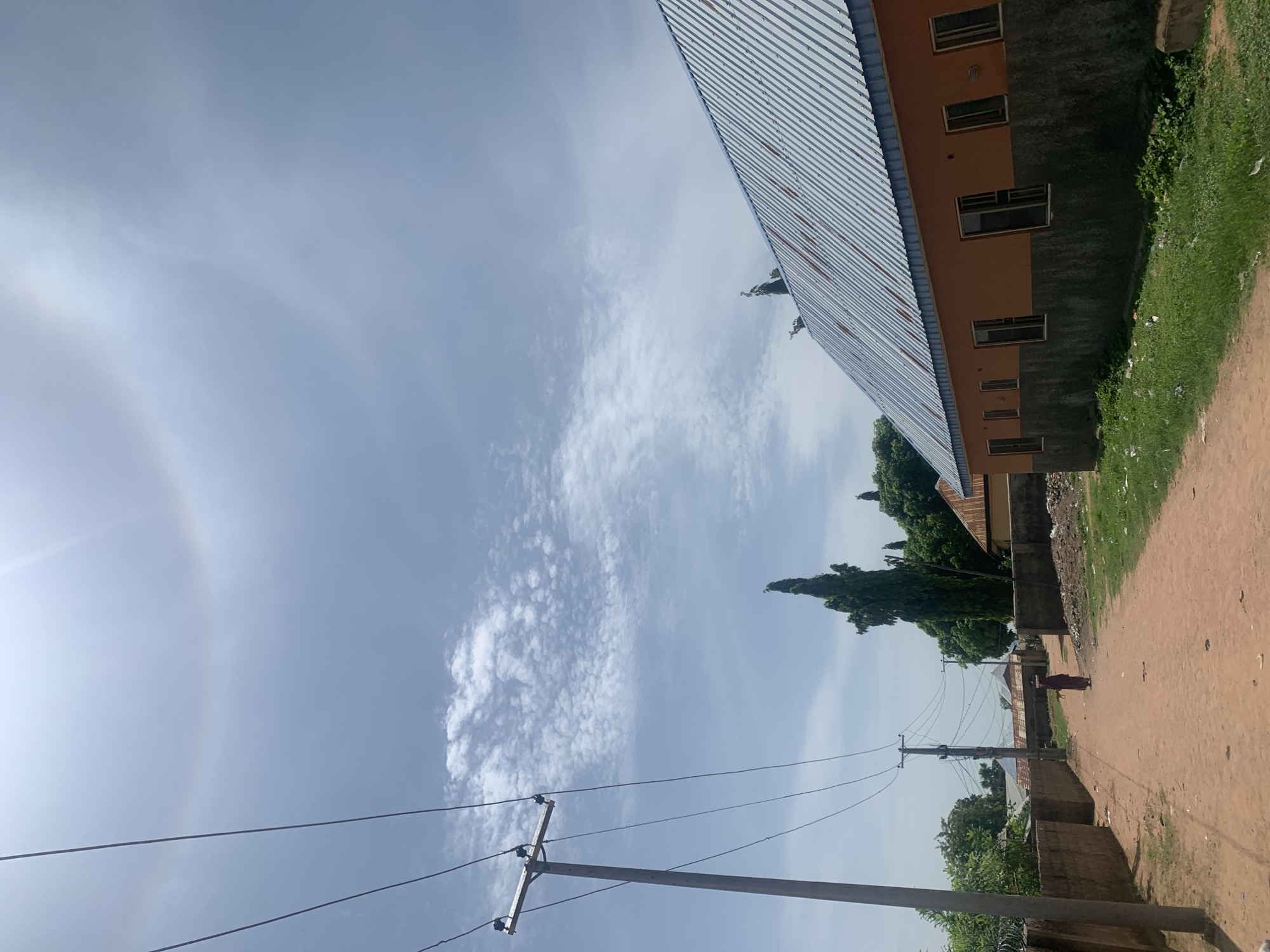 The image captures a sunlit scene on a dirt road in Jauro Yinu, Nigeria. A faint sun halo is visible in the sky, a phenomenon caused by ice crystals in the atmosphere refracting sunlight. Power lines stretch across the frame, supported by wooden utility poles. To the right, a single-story building with an orange upper wall and a grey textured lower wall has a corrugated metal roof. Several windows are visible on the building's facade. In the distance, a tall, slender evergreen tree stands prominently, surrounded by other foliage and buildings. A lone figure, presumably a woman in a dark garment carrying something on her head, walks away from the camera down the dirt road. The road is flanked by low brick walls on the left and a grassy verge with scattered debris on the right. The overall impression is of a rural or semi-urban setting under a bright sky.