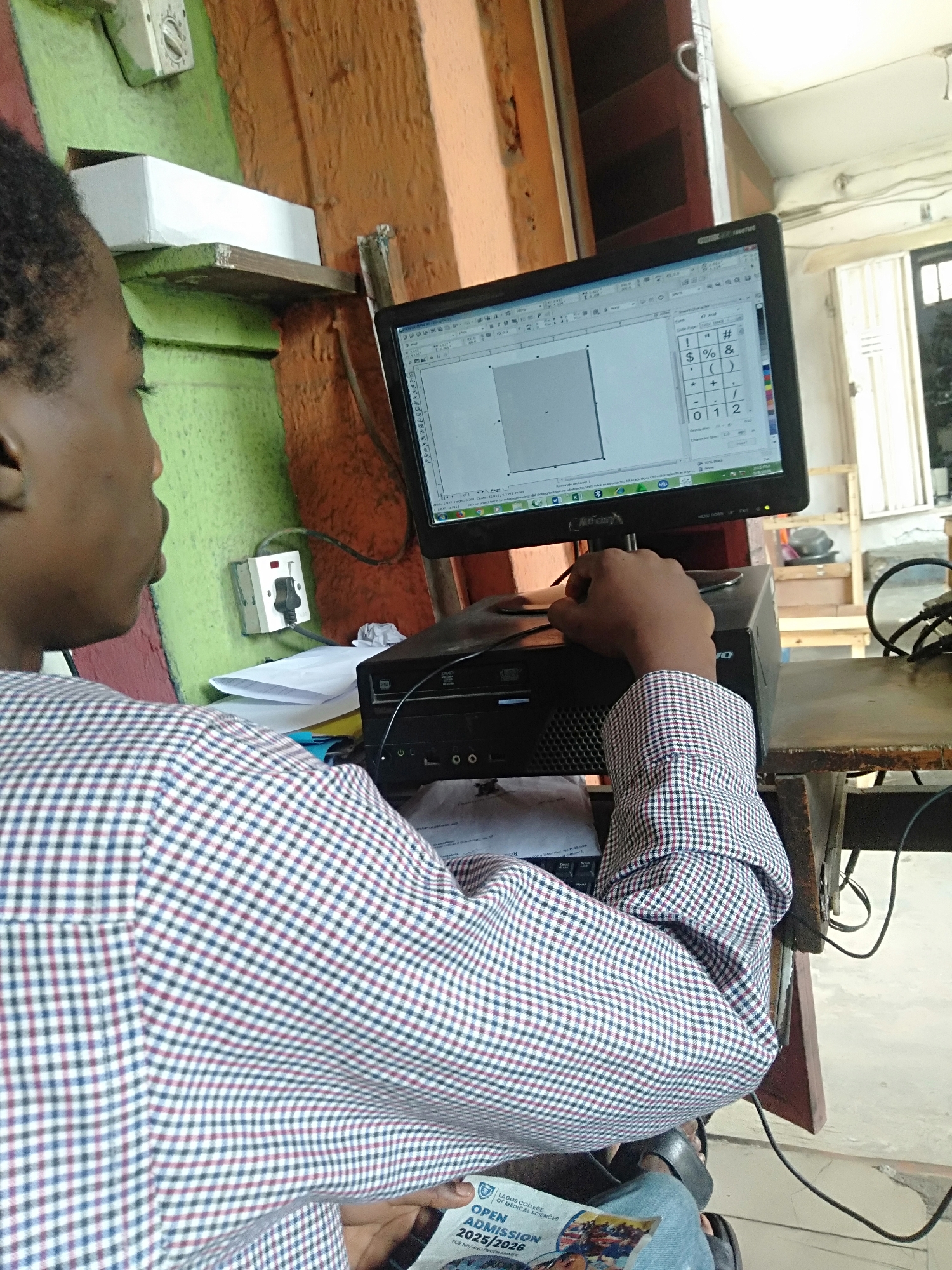 A person is sitting in front of a computer in a location that appears to be in Shangisha, Nigeria, based on the inscription "LAGOS COLLEGE OF MEDICAL SCIENCES" visible on a brochure held in their hands. The person is wearing a checkered long-sleeved shirt and blue jeans. They are in front of a desktop computer. The monitor displays a document with a grid and a character insertion menu. The CPU unit is black and located under the monitor. The environment appears to be an office or workspace with a wooden desk. The walls are painted in a combination of green and orange. Electrical outlets are visible on the wall. Cables and other objects are scattered around the desk.