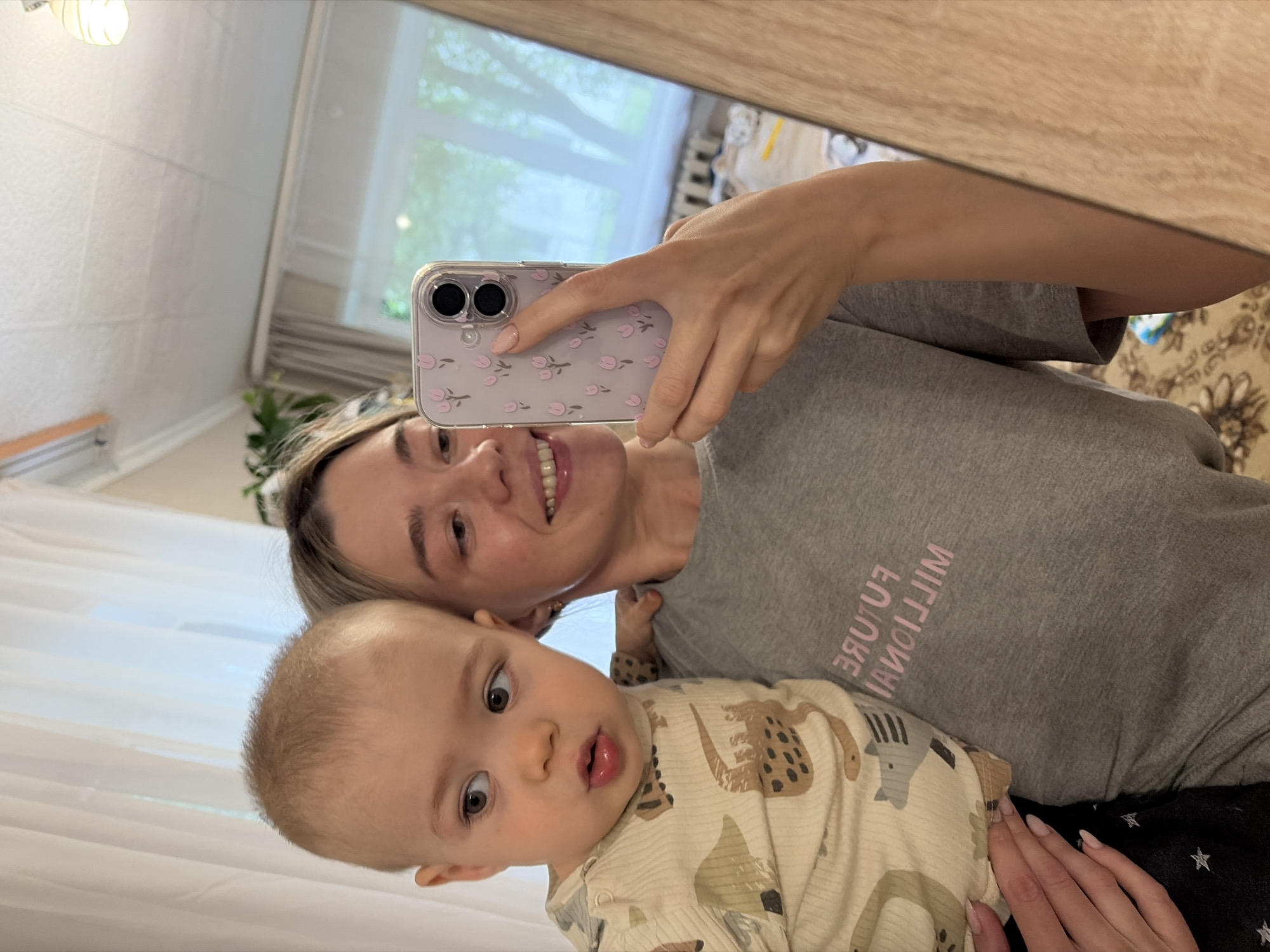 A selfie shows a woman and a baby. The woman is fair-skinned with blonde hair and is wearing a gray t-shirt with pink text that reads "FUTURE MILLIONAIR". She is holding a smartphone with a pale purple case decorated with small pink flowers. The baby is fair-skinned with light brown hair and is wearing a beige long-sleeved shirt with a pattern of animals. The background appears to be an interior space, possibly a home, with curtains, a window, and furniture visible in the reflection of the mirror. It is not possible to confirm the location of this image to be Chaikovsky, Russia.
